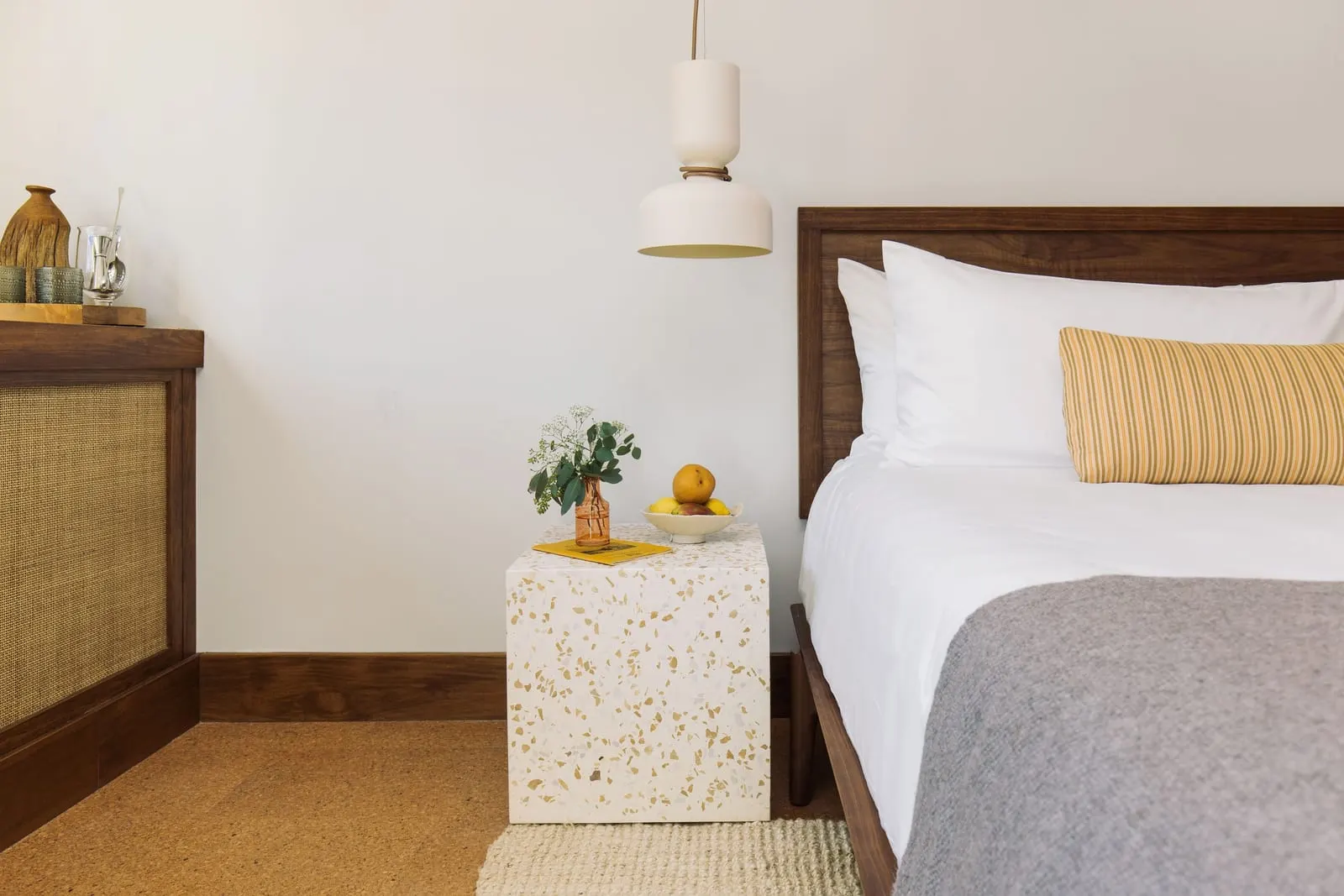 The Queen at Silver Lake Pool & Inn: a bed with white linen and a grey throw beside a terrazzo bedside table with a fruit bowl, under a pendant lamp