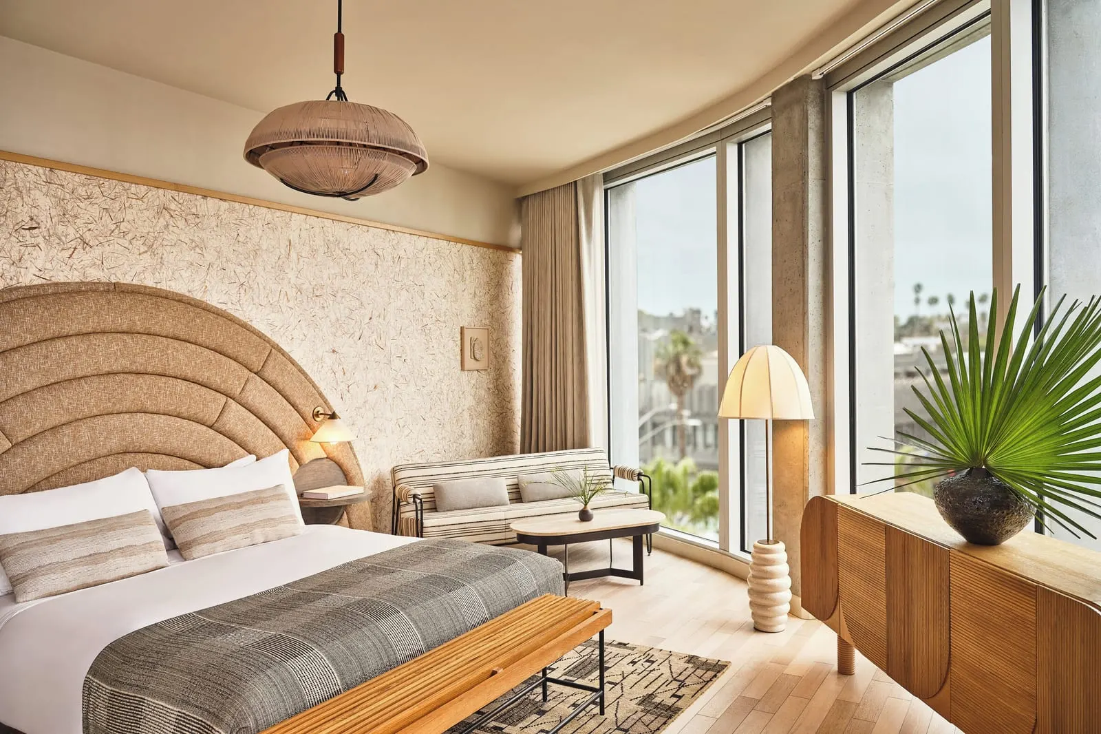 Deluxe Suite im Santa Monica Proper Hotel: ein Kingsize-Bett mit einer Bank am Fußende, ein gestreiftes Sofa und ein niedriger Tisch vor einer geschwungenen Wand aus raumhohen Fenstern