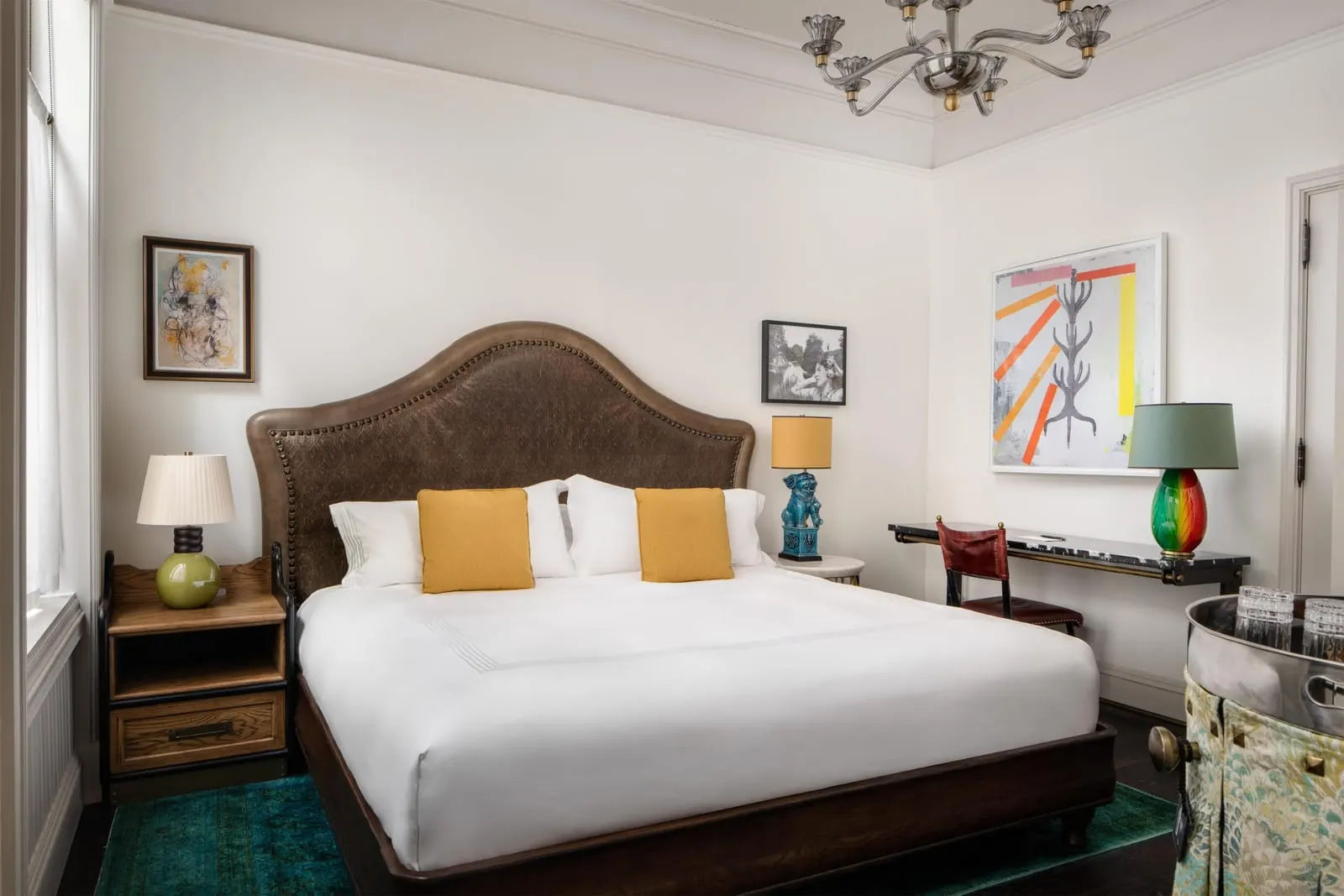 Superior King at The Beekman: a king bed with a studded leather headboard and yellow cushions, a narrow marble desk under abstract art and a skirted drinks cart at the right