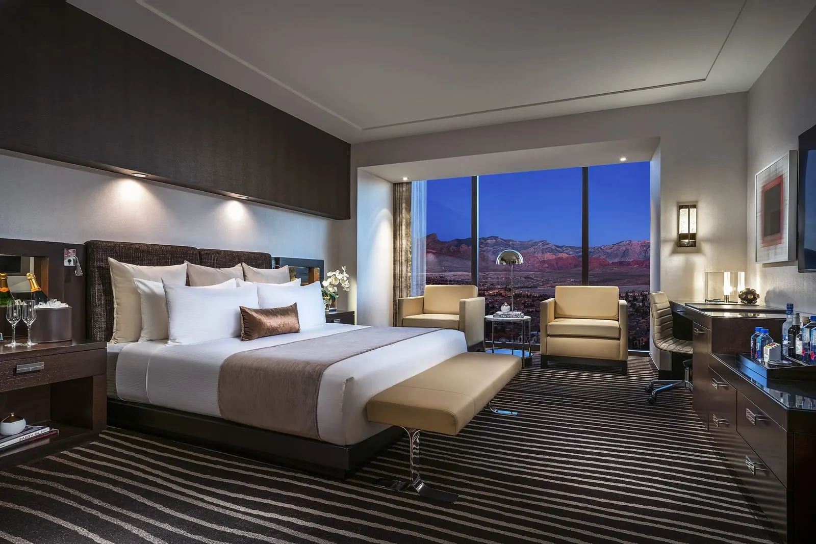 Canyon View King at Red Rock Casino Resort & Spa: a king bed with an upholstered bench at its foot, two armchairs by a floor-to-ceiling window, and a desk and dresser along the right-hand wall