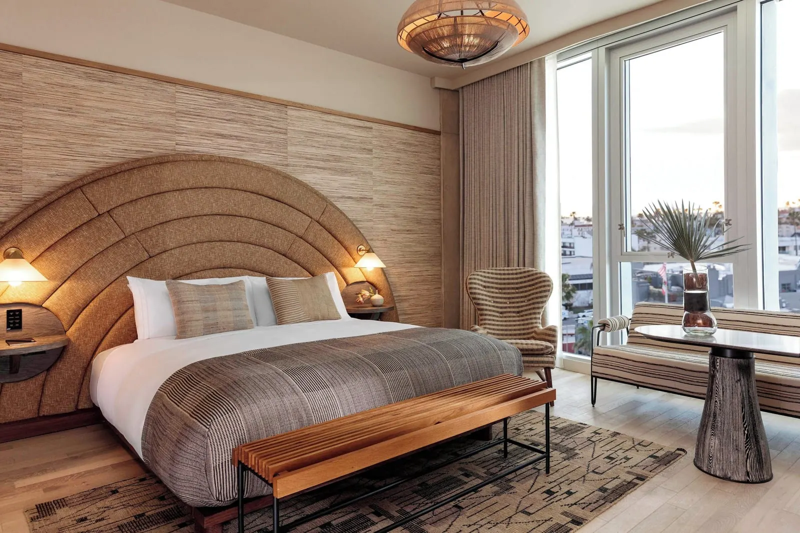 Premier King im Santa Monica Proper Hotel: ein Kingsize-Bett auf einem Webteppich mit einer Holzlattenbank am Fußende, und ein Sofa und ein Tisch am raumhohen Fenster