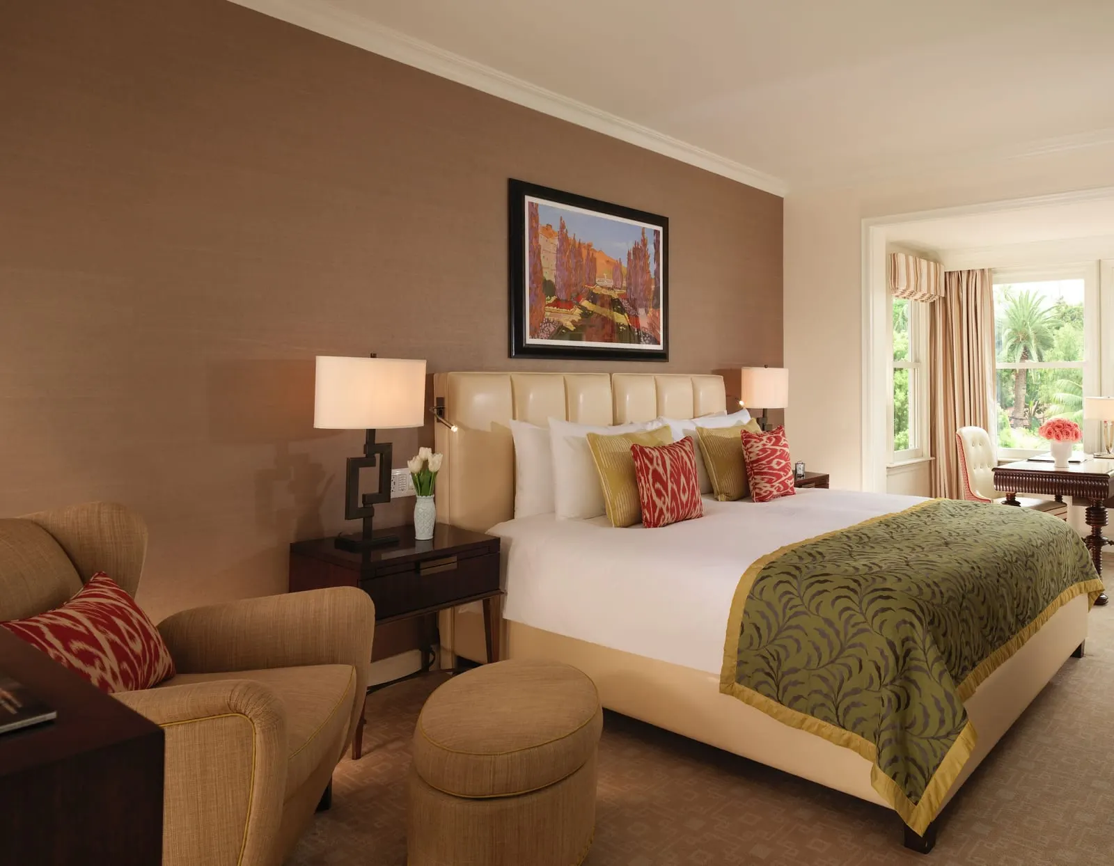 Superior Room with Balcony at The Beverly Hills Hotel: a bed with a quilted cream headboard and a green leaf-patterned runner, a tan wing armchair with a footstool and a writing desk in a bay window