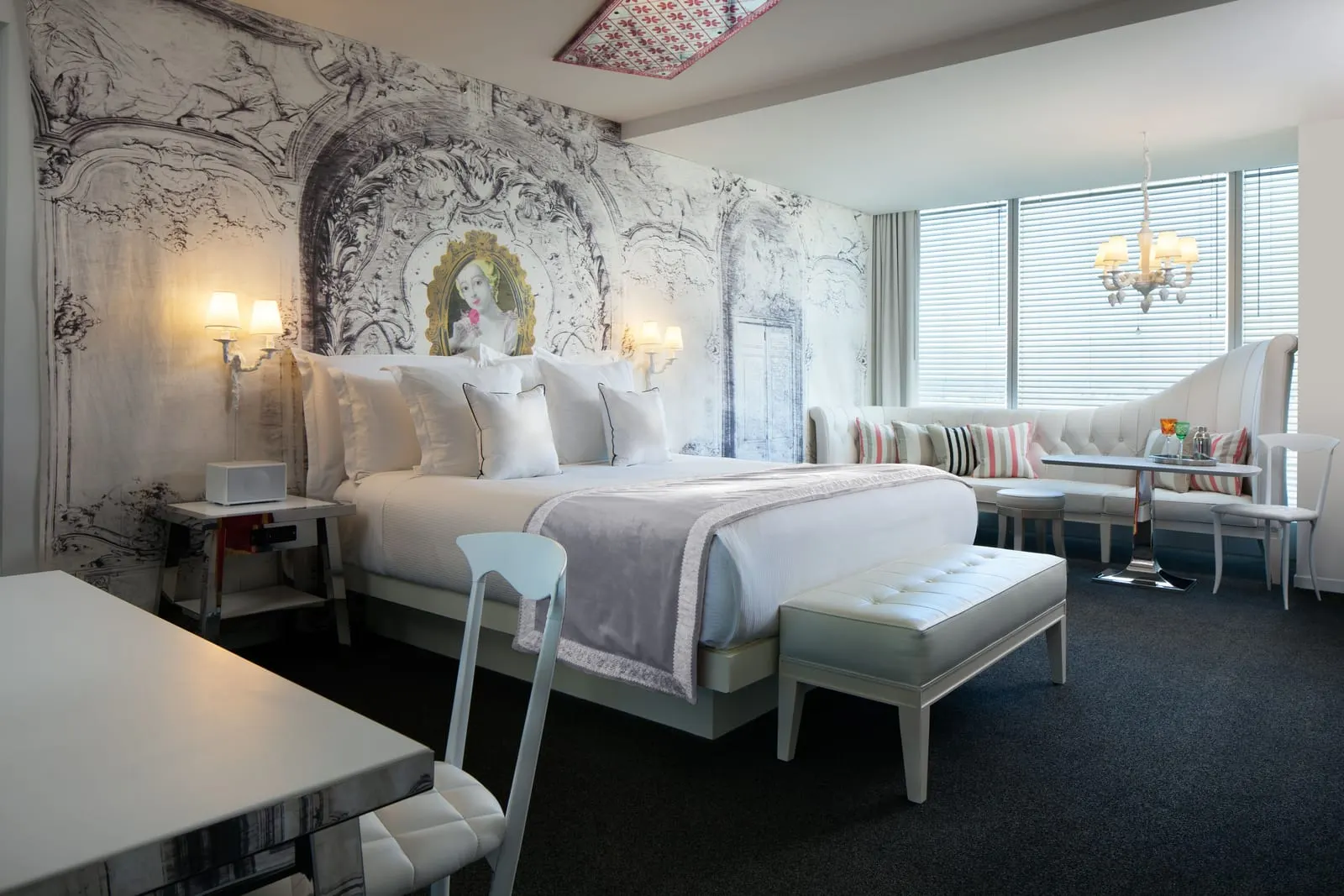 Alexandria Ultra Suite at SAHARA Las Vegas: the bedroom, with a king bed against a black and white baroque-print wall with a framed portrait, a bench at its foot, and a white tufted banquette and table by a wide window