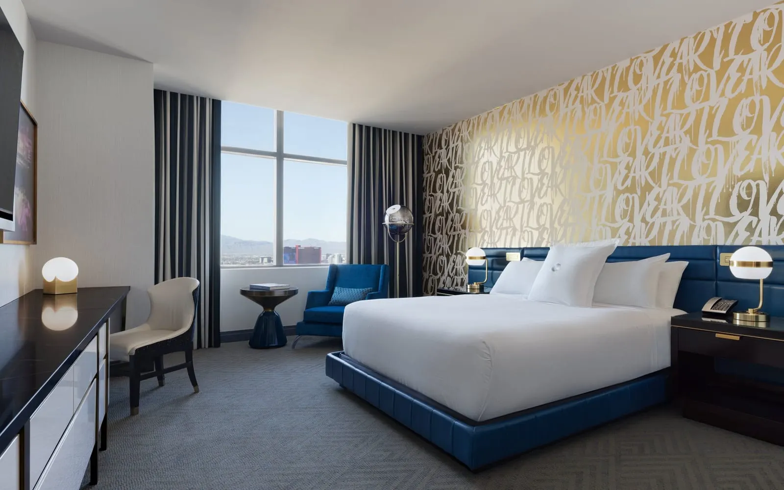 City Room at The Cosmopolitan of Las Vegas: a king bed on a blue leather base against a gold wall painted with white lettering, a blue armchair by the window and a desk with a cream chair