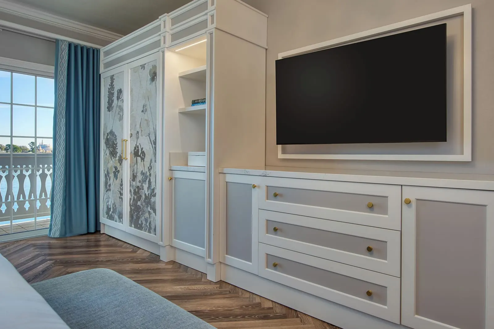 1 BR - Theme Park View - Club Level Access suite at Disney's Grand Floridian Resort & Spa: a wardrobe with painted floral doors, a wall-mounted television above a white chest of drawers and a glass door onto a balcony with white railings