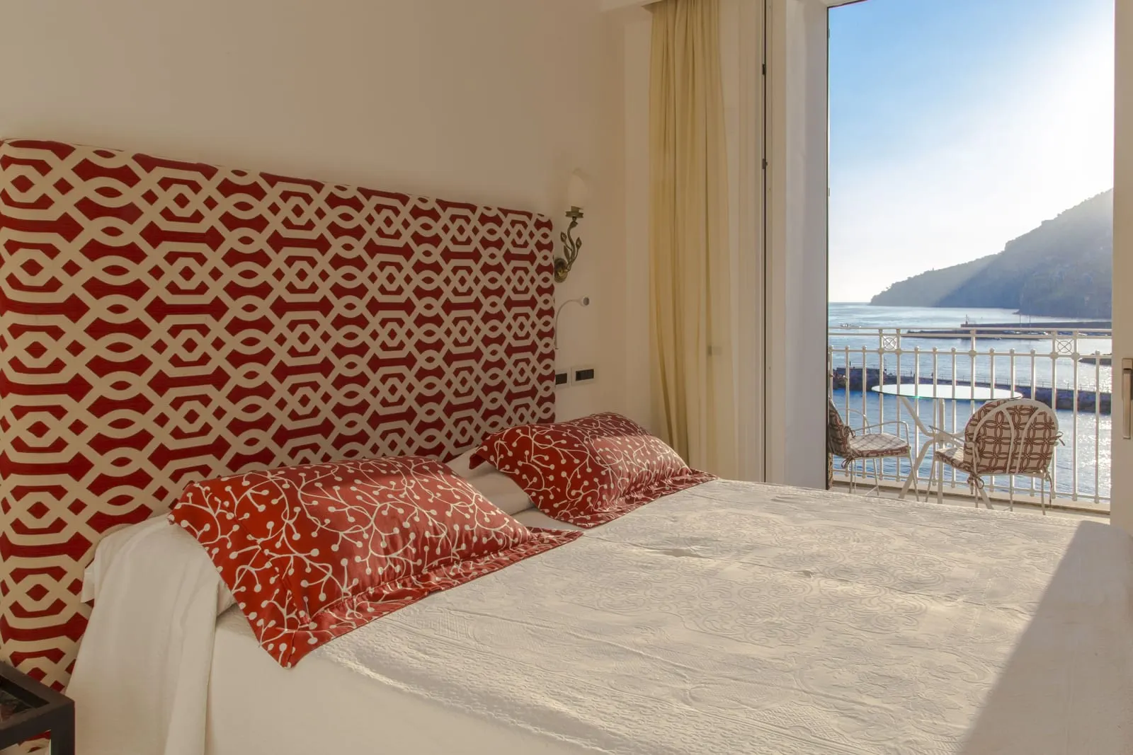 Deluxe at Hotel Marina Riviera: a double bed with a red and white headboard beside an open door to a balcony with two chairs over the sea