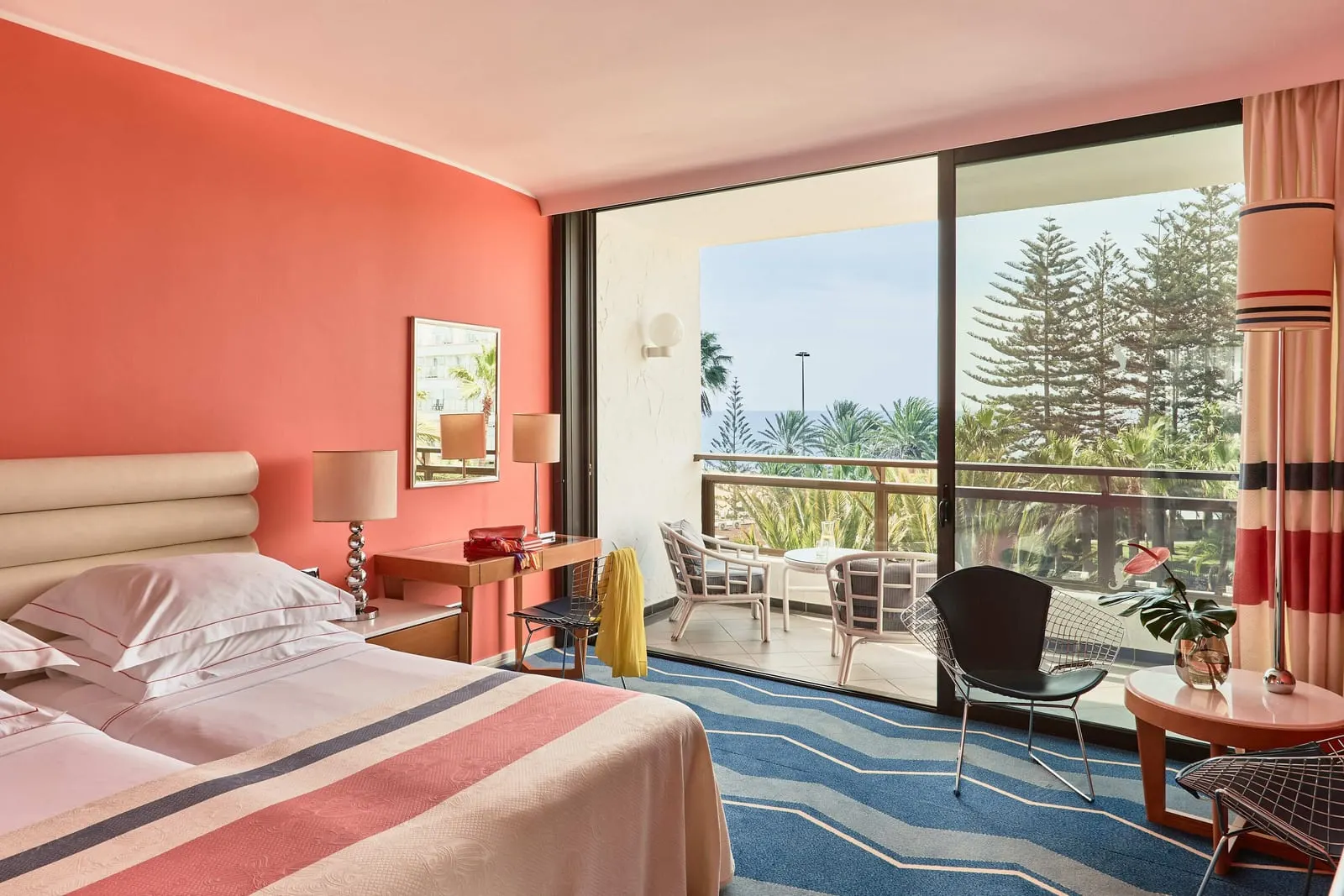 Superior Room at Seaside Palm Beach: twin beds against a coral wall, a blue zigzag carpet and a sliding door to a balcony with cane chairs