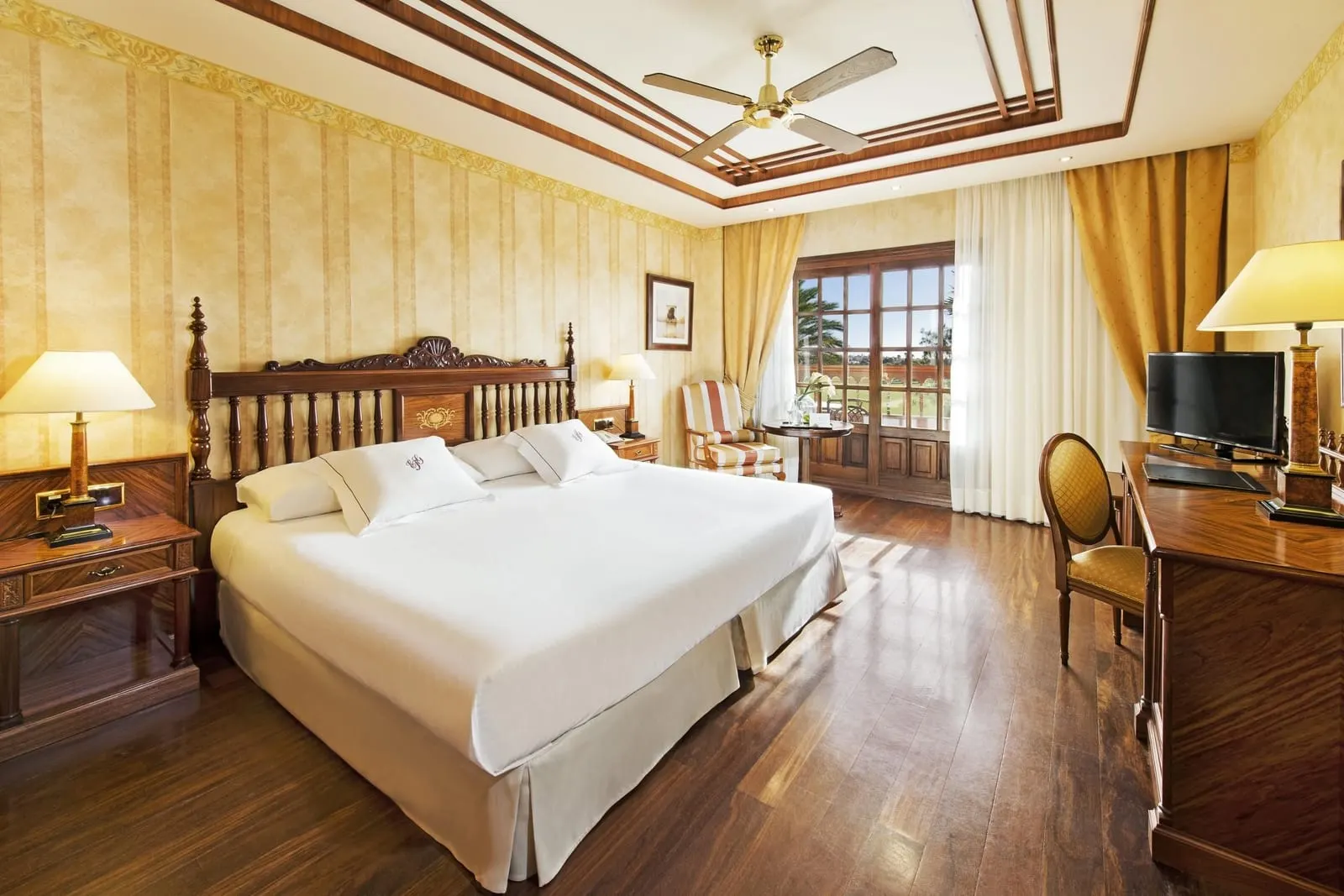 Deluxe double room at Elba Palace Golf Boutique Hotel: a king bed with a carved wooden headboard, wooden floors, a desk with a television and a ceiling fan