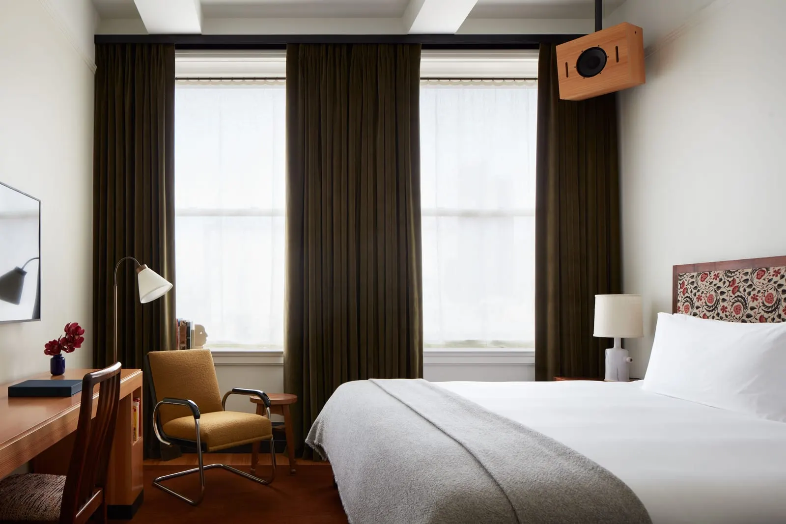 Studio King at Nine Orchard: a bed with a grey wool throw in front of two oversized windows with long dark curtains, a yellow armchair and a wooden desk at the left and a wall speaker above