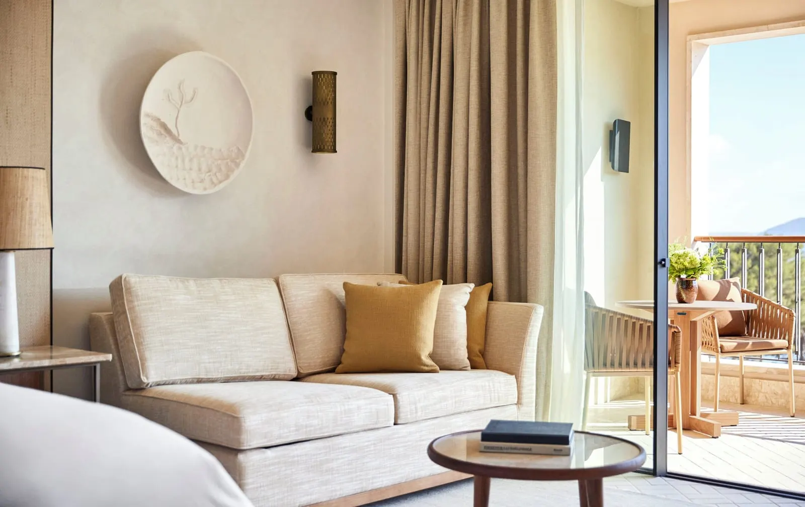 Grand Sea-View Room im Four Seasons Resort Mallorca at Formentor: ein cremefarbenes Sofa mit senffarbenen Kissen unter einem runden Gipsrelief, neben der Ecke des Bettes und einer offenen Tür zur Terrasse