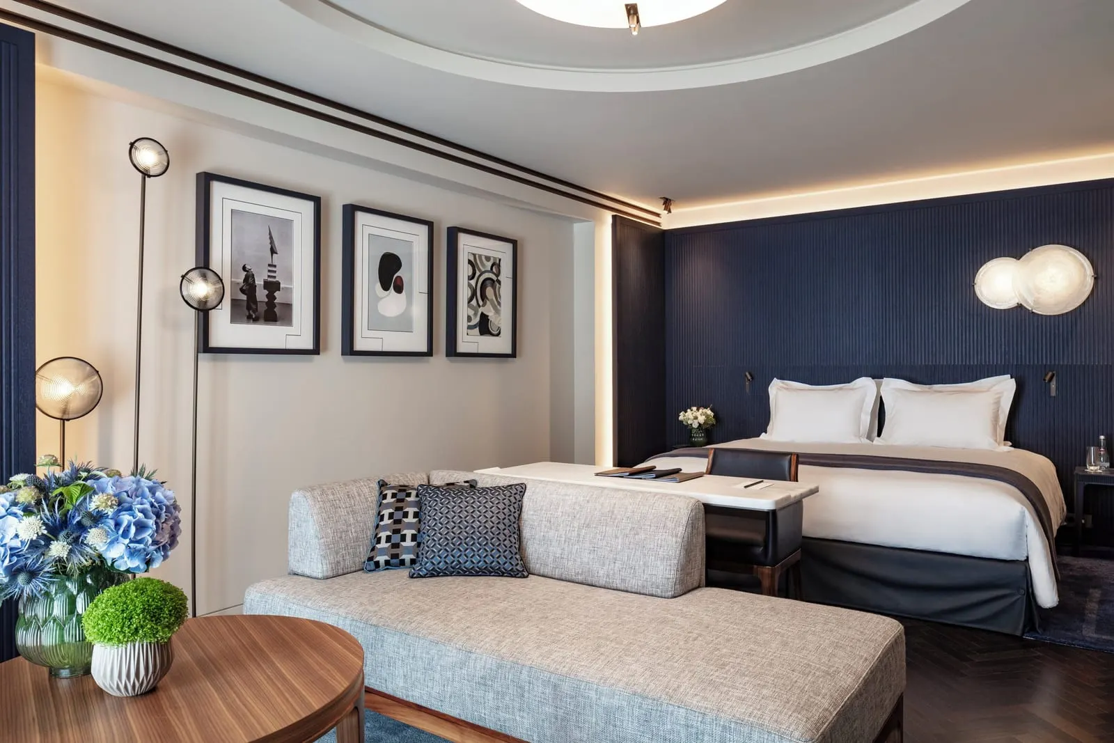 Junior Suite Balcony at Mandarin Oriental Lutetia, Paris: a double bed against a dark blue fluted headboard, with a desk at its foot and a grey sofa, a round table and three framed prints in the lounge area