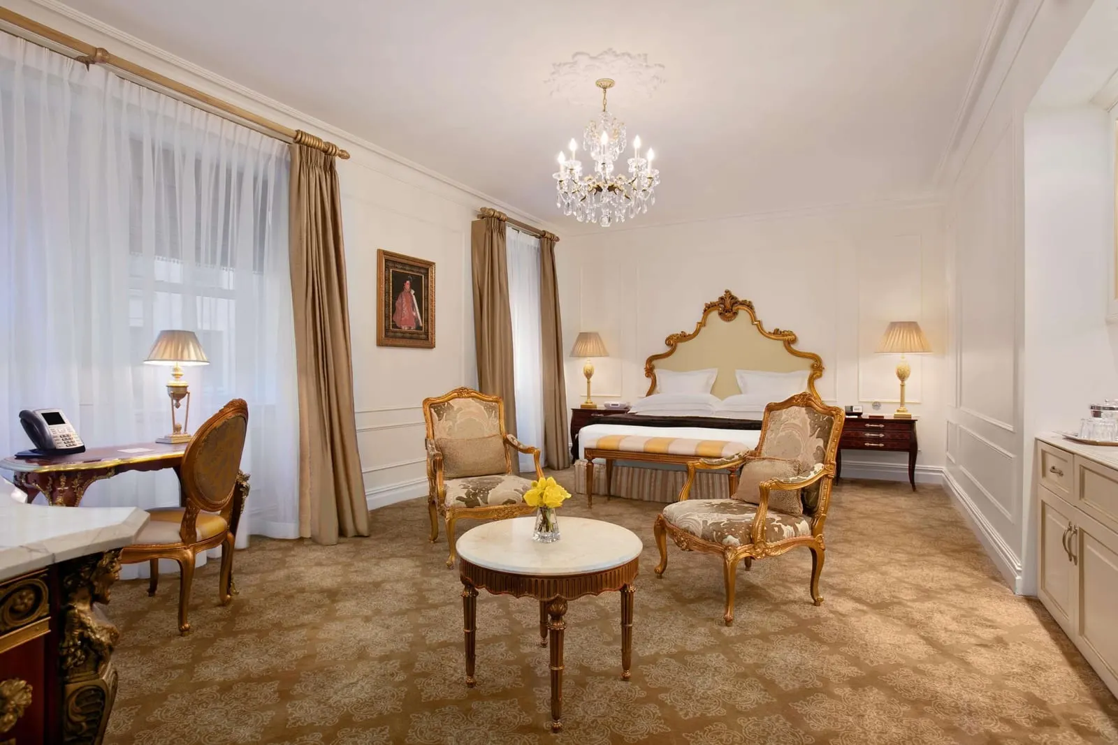 Grand Luxe King Room at The Plaza: an open plan room with a king bed at the far end, two gilt armchairs around a marble table, a writing desk and a pantry cabinet along the right wall
