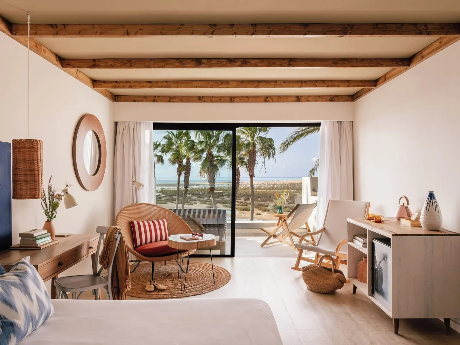 Zel Junior Suite Ocean View at ZEL Fuerteventura: a sitting corner with a wicker armchair and a rocking chair by a glass door, with palm trees and the sea outside