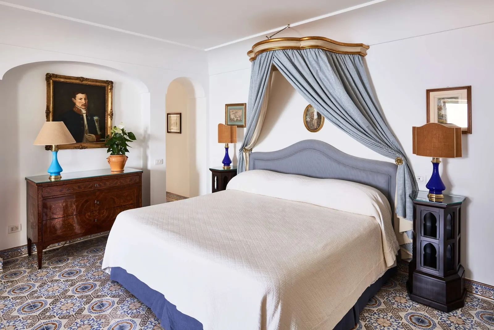 One Bedroom Suite Sea View at Le Sirenuse: a double bed under a blue canopy, with an inlaid chest of drawers and a portrait above it