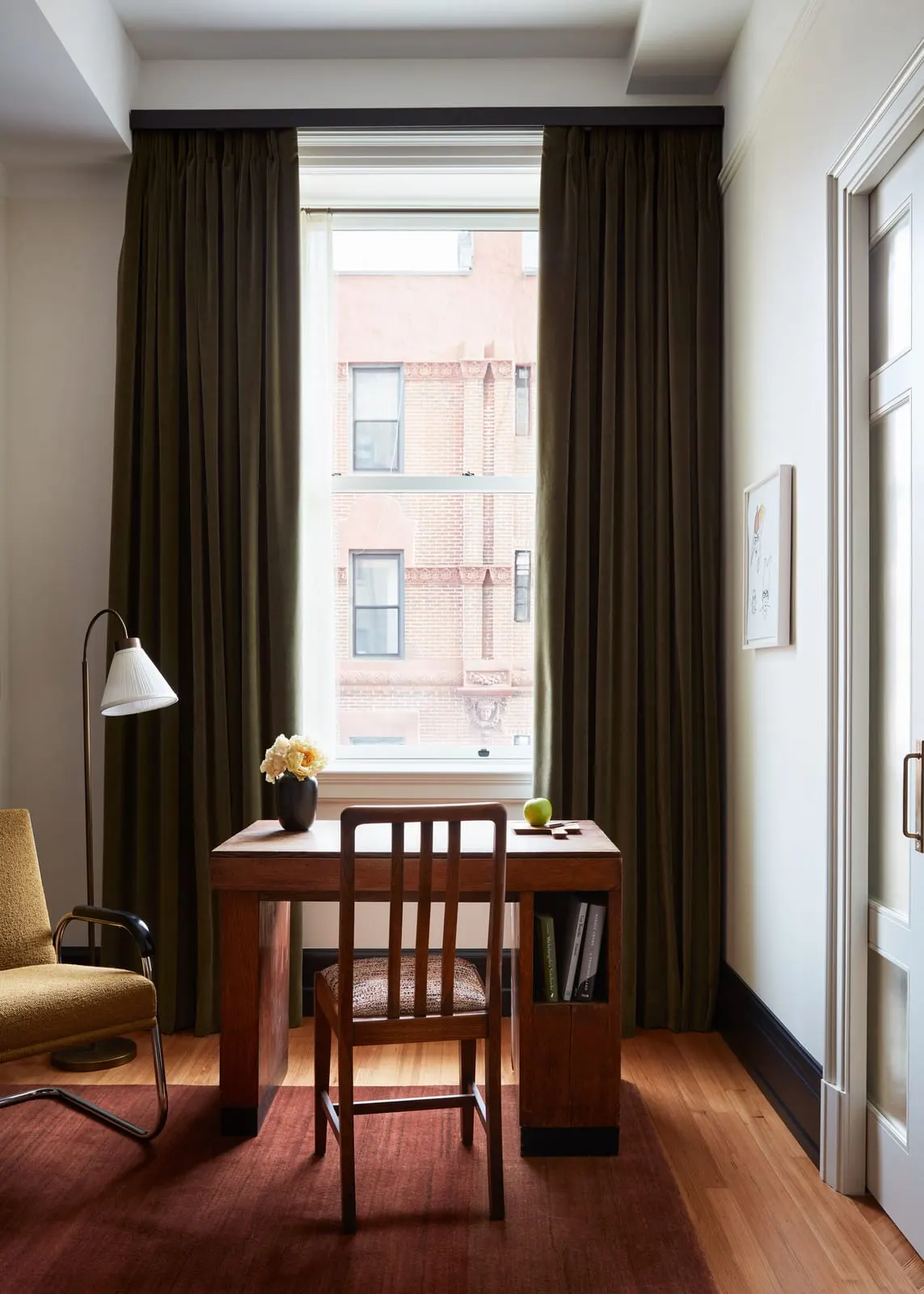 Corner King at Nine Orchard: a wooden desk and chair in front of a tall window with long olive curtains, a floor lamp and a yellow armchair to one side