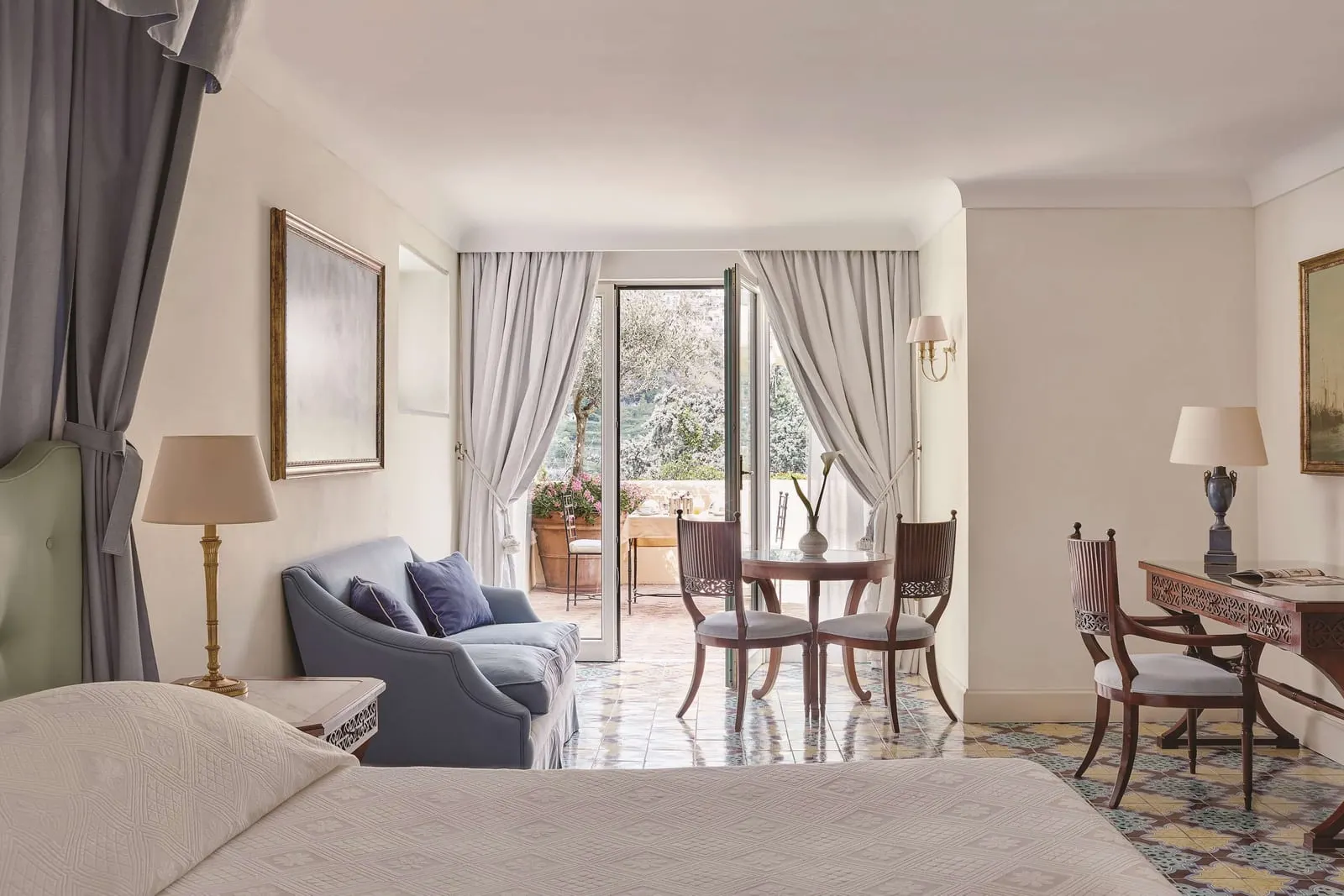 Garden View Room with Terrace at Caruso, A Belmond Hotel: a bed, a blue sofa, a desk and a small dining table on a tiled floor, with doors open onto a terrace with potted flowers and trees