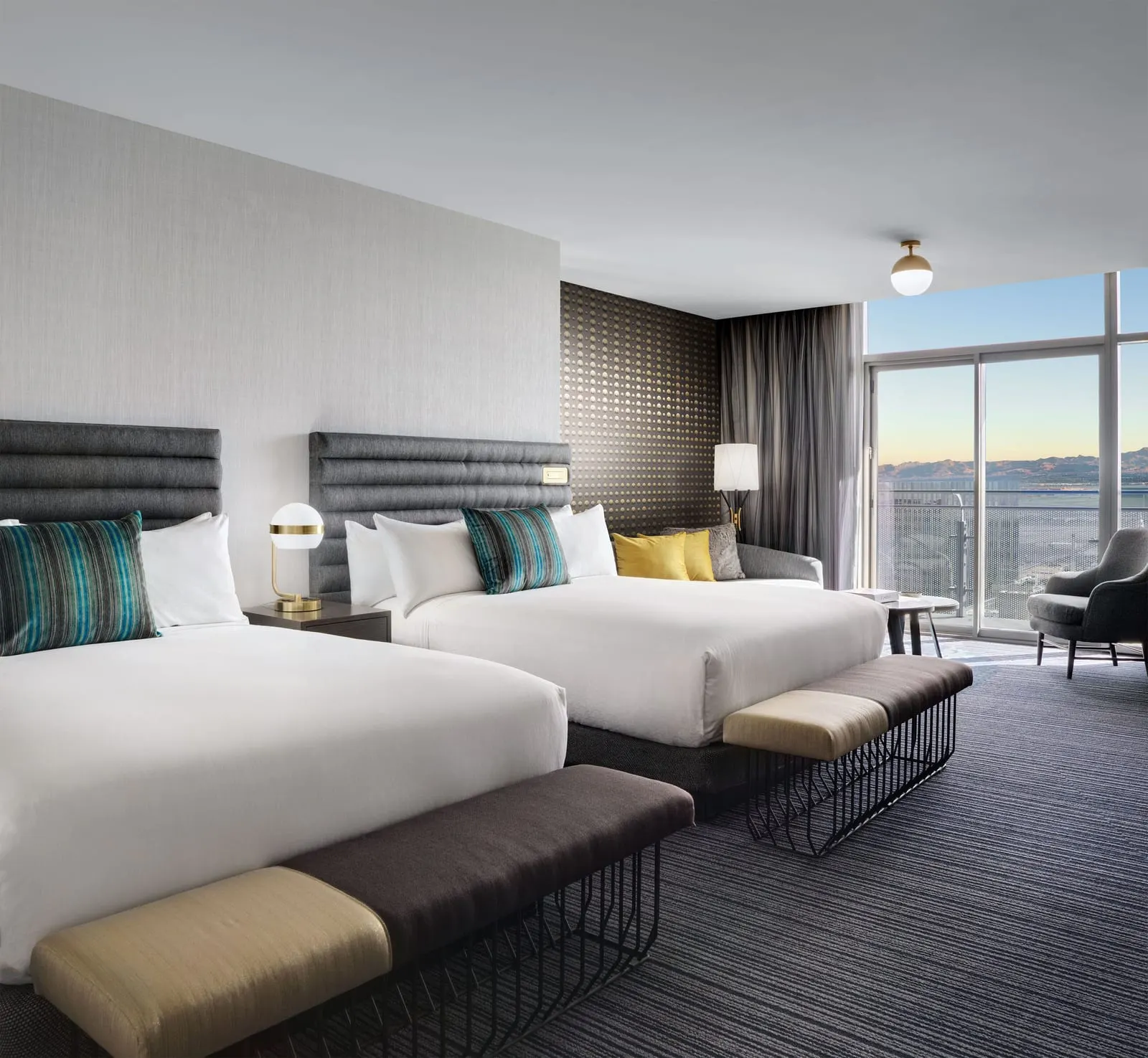 Terrace Studio Two Queen at The Cosmopolitan of Las Vegas: two queen beds with padded grey headboards and benches at their feet, a sofa with yellow cushions and an armchair by full-height glass doors
