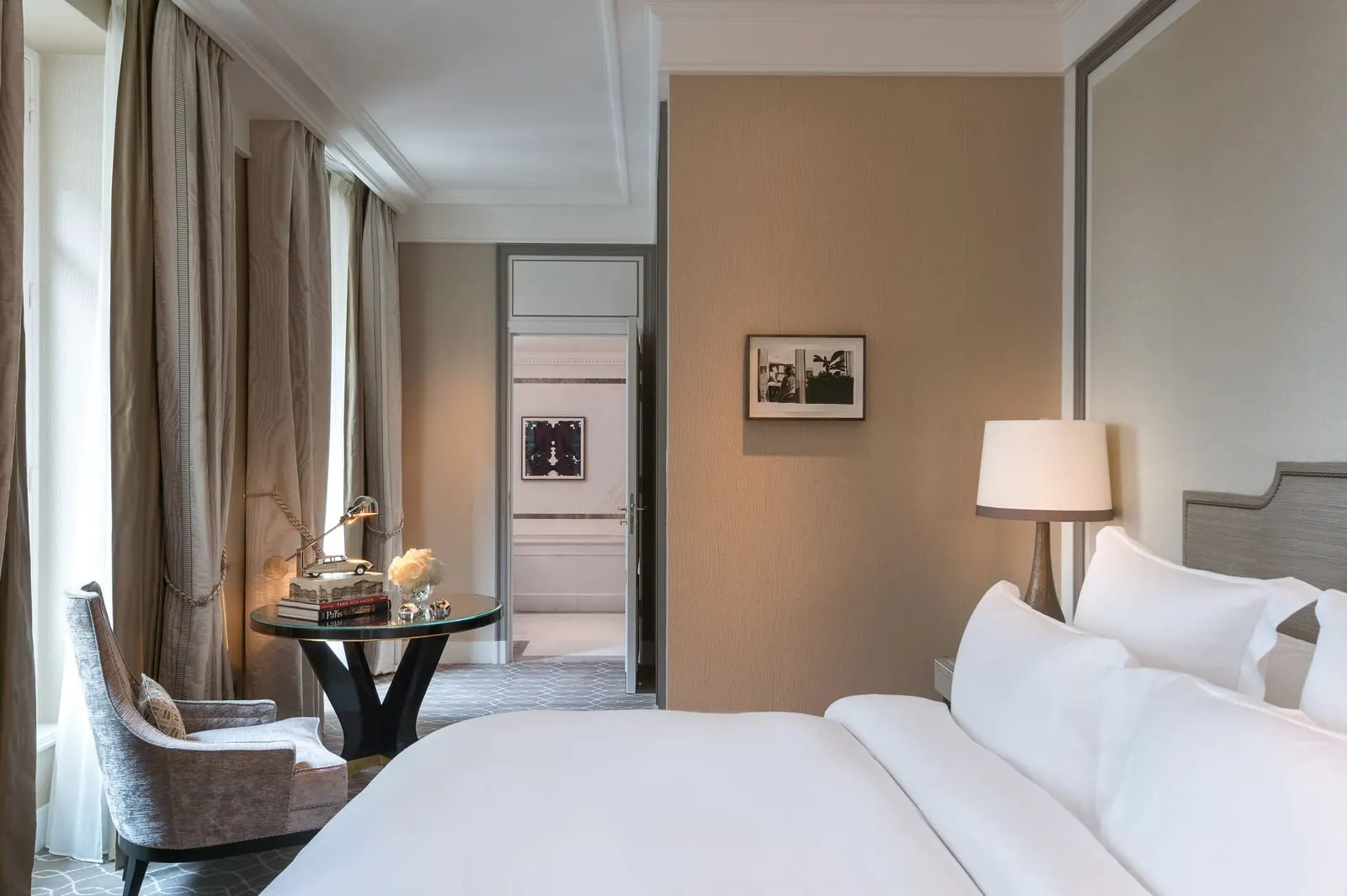 Deluxe Room at Hôtel de Crillon, A Rosewood Hotel: the bed with an upholstered headboard, a round glass table and a velvet armchair by tall curtained windows, and an open door through to the bathroom
