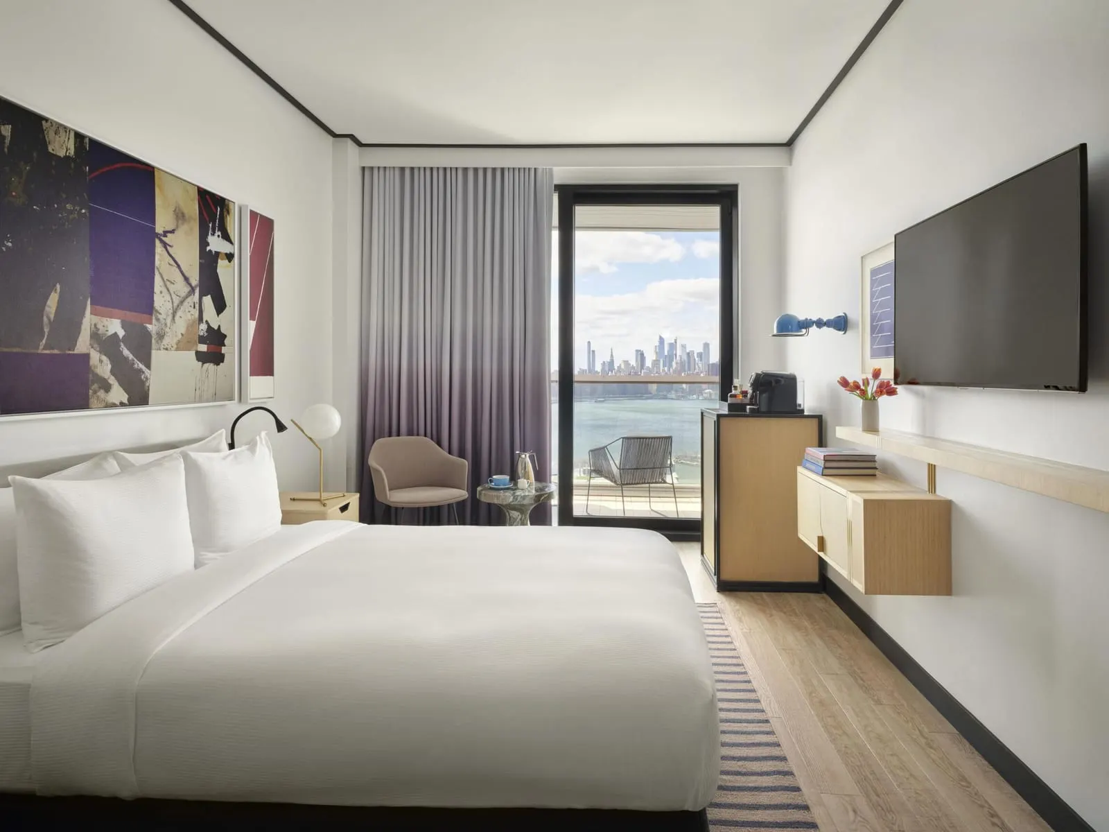 Manhattan View Queen with Balcony at The William Vale: a bed with white linen under two large abstract paintings, a wall-mounted television, and a glass balcony door framing a chair, the river and the Manhattan skyline