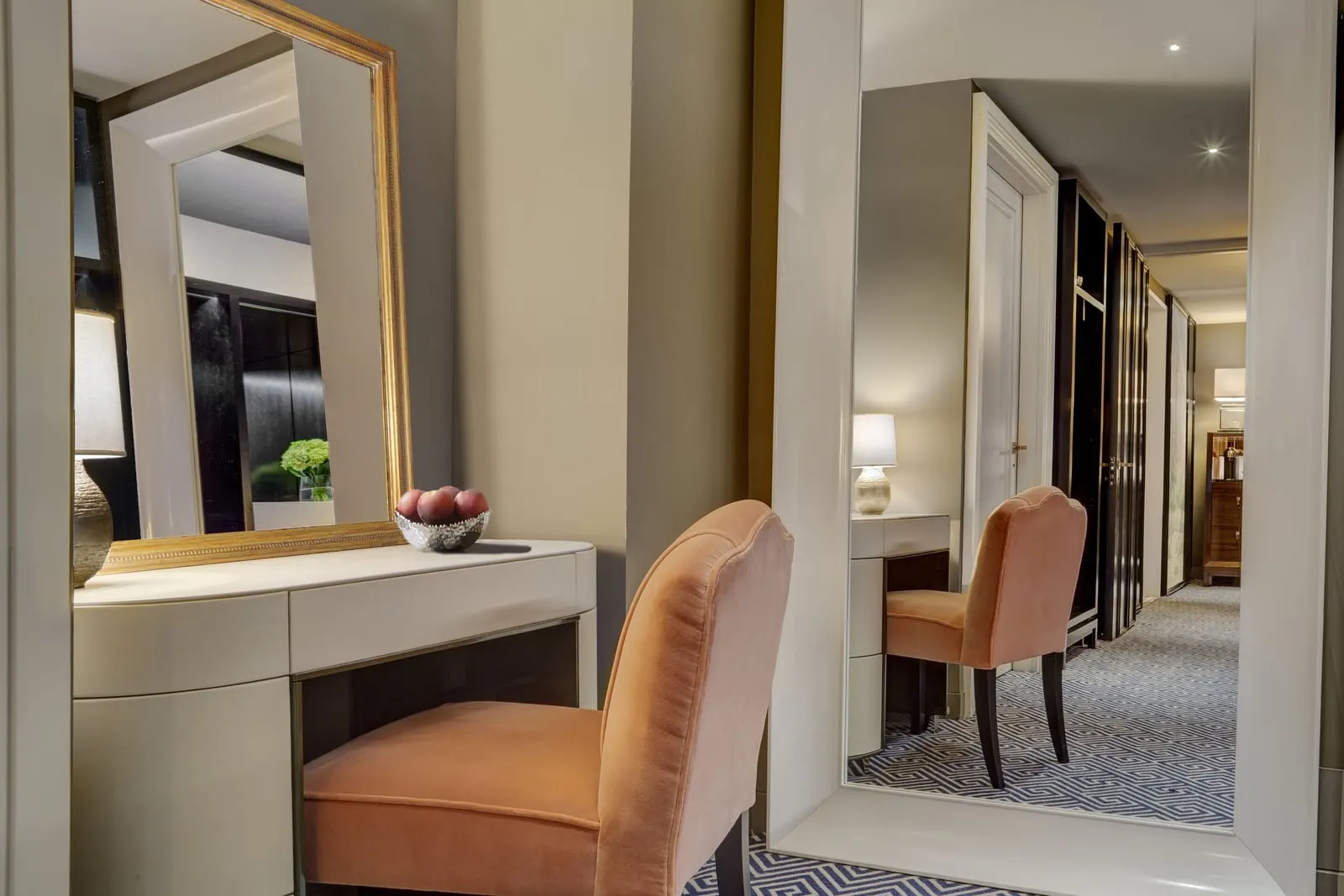 Premier Room at The Carlyle: a dressing table with a gold-framed mirror and a peach velvet chair, with a tall mirror reflecting the hallway