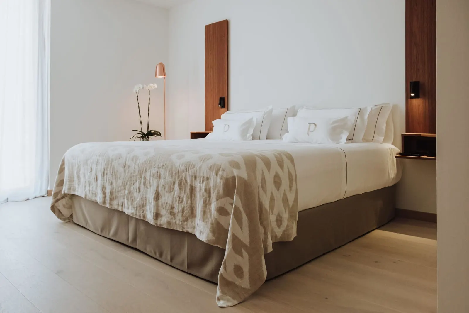 Signature Palma Sea View Suite at Es Princep: a wide bed with monogrammed pillows and a woven bedspread, beside a copper floor lamp and an orchid