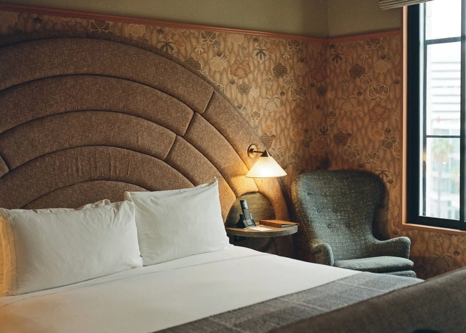 Proper Sleep Room at Santa Monica Proper Hotel: a bed with a tall arched headboard, a bedside table with a lamp and a grey armchair by a window, against patterned wallpaper