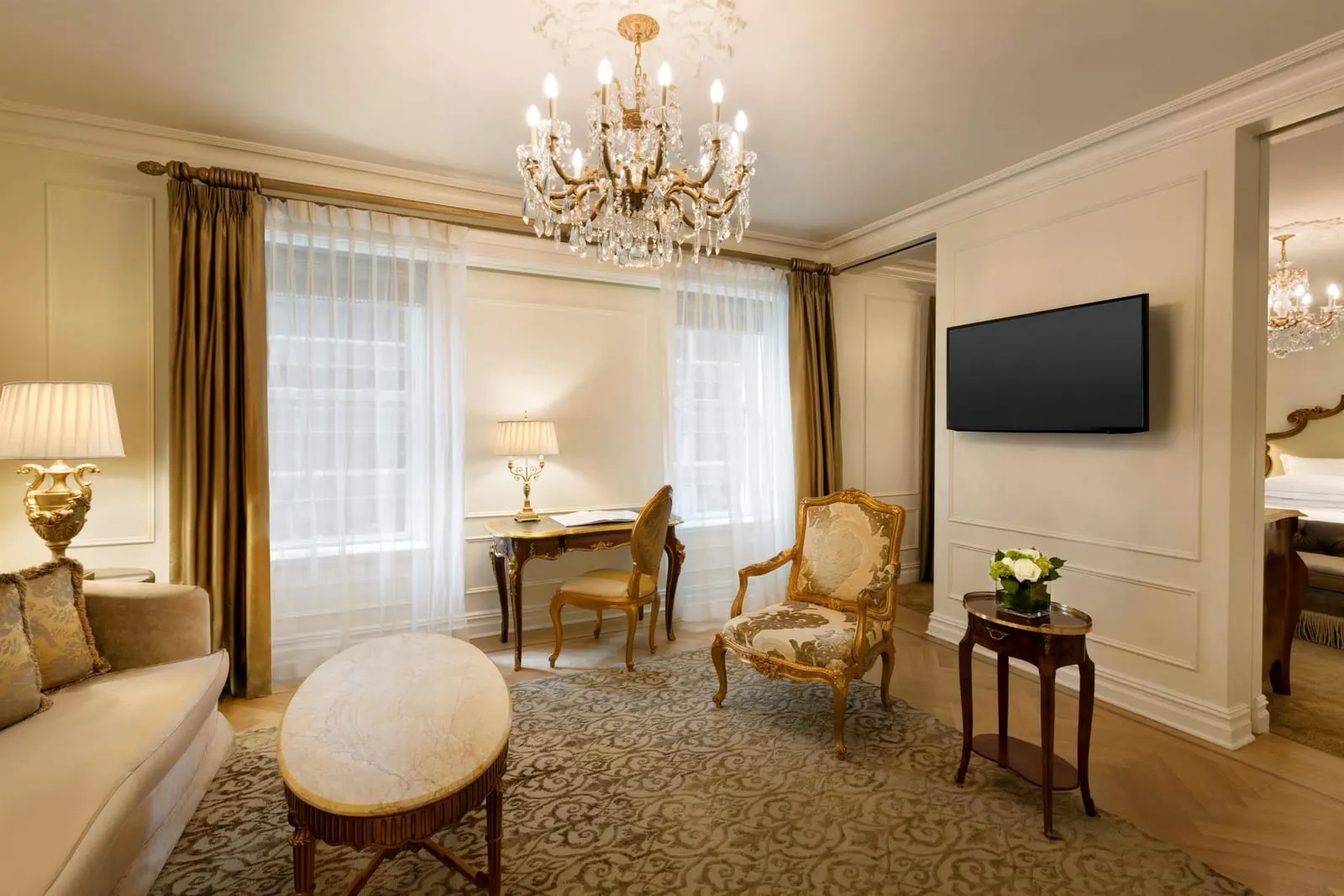 Junior Suite King at The Plaza: a sitting room with a sofa, a marble coffee table, a writing desk and a gilt armchair, and the bedroom visible through an opening on the right