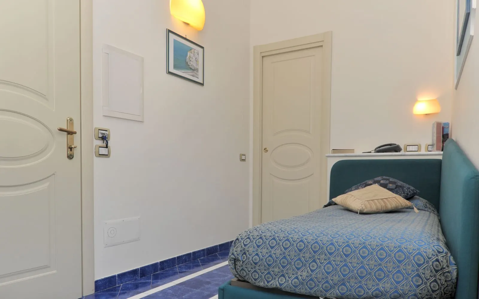 Economy Single at Hotel Santa Lucia: a single bed with a teal frame and a blue patterned cover beside the entrance door