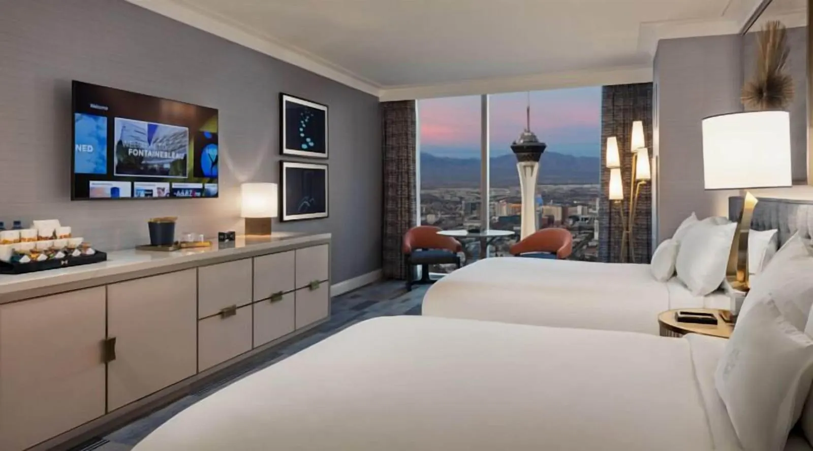 Gold Queen Mountain Great View at Fontainebleau Las Vegas: two queen beds, a long cabinet under a wall television, and two chairs at a round table by floor-to-ceiling windows onto the city and the mountains at dusk