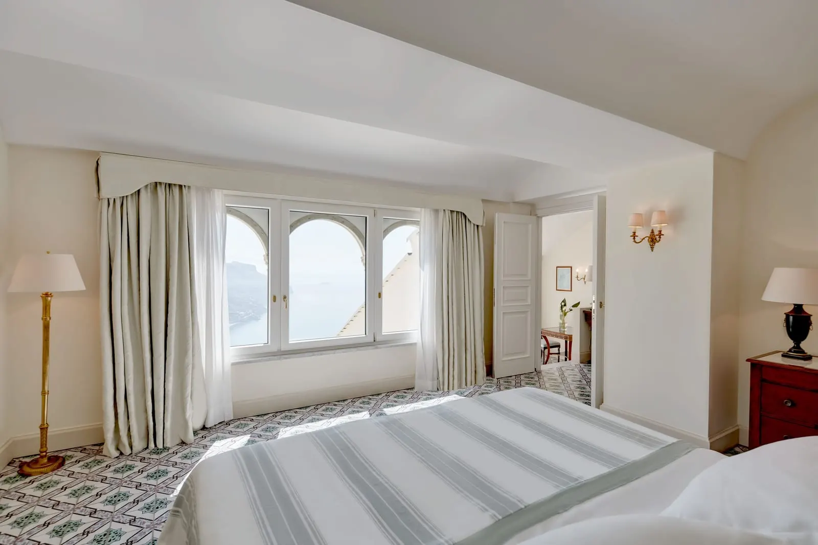 Deluxe Sea View Terrace Suite at Caruso, A Belmond Hotel: a bed facing arched windows over the sea and the coast, with a door through to the living room