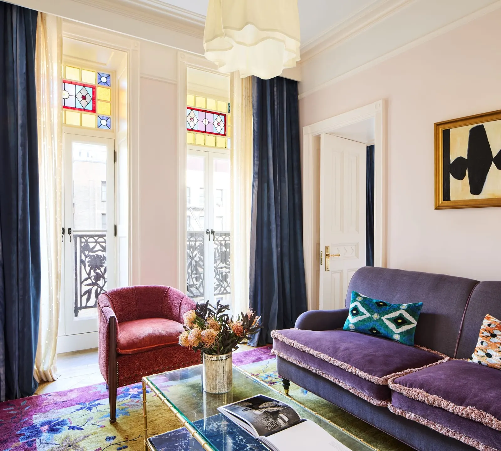 Petite Suite at Hotel Chelsea: a sitting room with a purple sofa, a rust-red armchair and a glass coffee table, in front of two tall glazed doors with stained-glass panels above and an iron railing outside