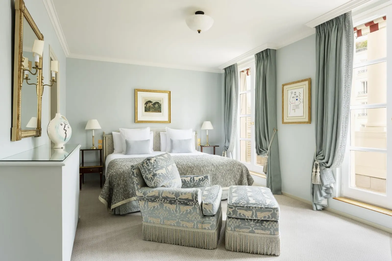 Suite Azur at Le Bristol Paris: a bed with a patterned grey throw, an armchair and footstool in matching fabric, and tall windows with grey-blue curtains