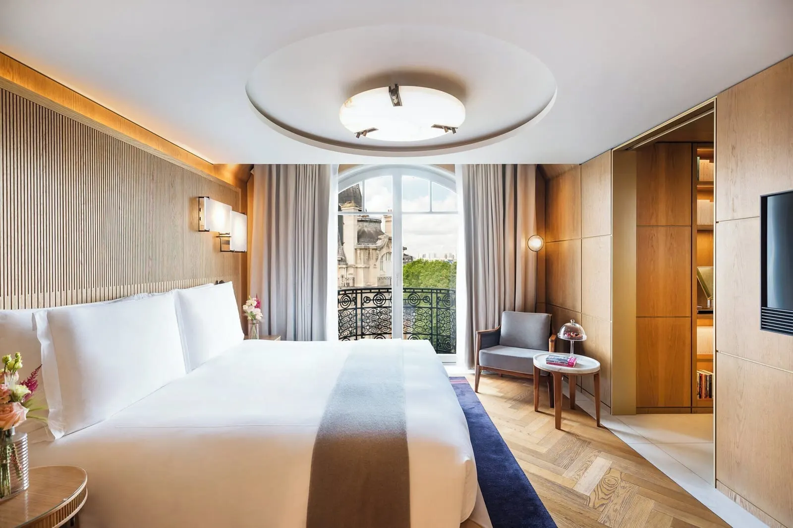 Eiffel Suite at Mandarin Oriental Lutetia, Paris: a double bed against an oak-panelled wall, an armchair and side table, and an arched French window onto a wrought-iron balcony, with bookshelves through the open door