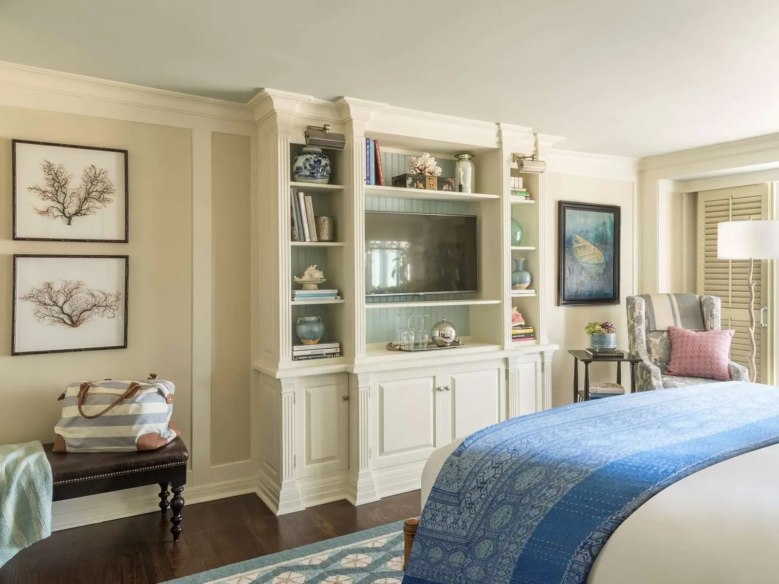 Partial Ocean View Room im Shutters on the Beach: ein cremefarbener Einbauschrank mit Fernseher und Regalen, zwei gerahmte Korallendrucke, eine Lederbank und das Fußende eines Bettes mit einer blau gemusterten Tagesdecke