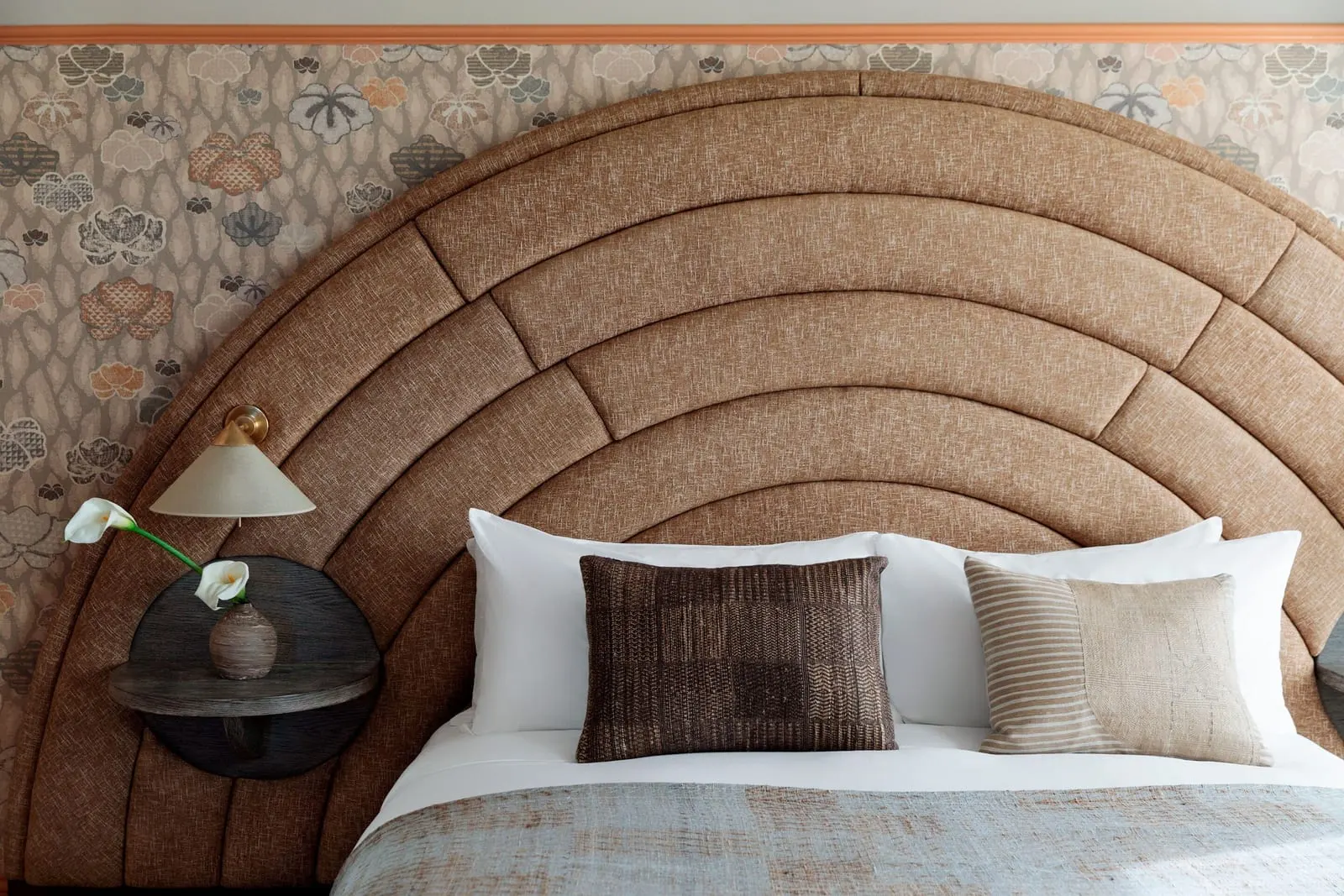 Landmark Deluxe Queen im Santa Monica Proper Hotel: eine Nahaufnahme eines Bettes mit einem hohen gerillten bogenförmigen Kopfteil, einer Wandlampe und einem runden Nachttischregal vor geblümter Tapete
