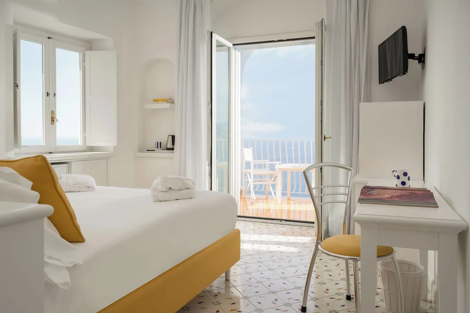 Camera Comfort with sea view balcony all'Hotel Tramonto d'Oro: un letto matrimoniale su un pavimento piastrellato dipinto a mano, con una scrivania e porte aperte su un balcone con tavolo, sedie e il mare in lontananza.