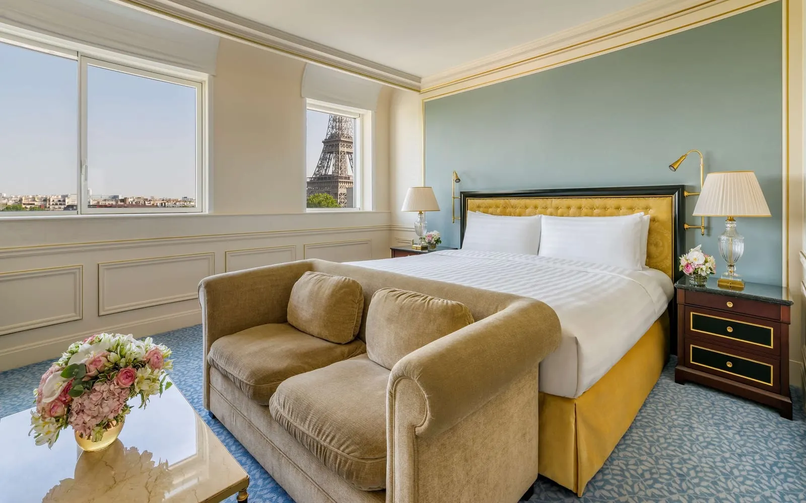 Terrace Eiffel View Room at Shangri-La Paris: a king bed with a gold headboard against a blue wall and a velvet sofa at its foot, with the Eiffel Tower in a side window