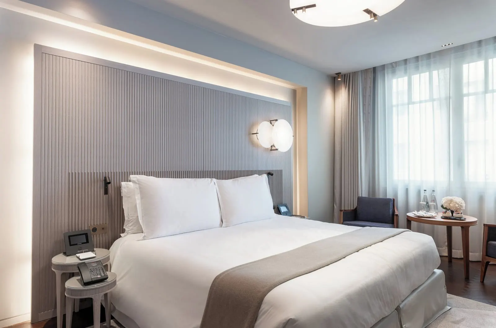Family Deluxe Suite at Mandarin Oriental Lutetia, Paris: a double bed against a grey fluted headboard with two round glass wall lights, and an armchair and a round table by windows with sheer curtains
