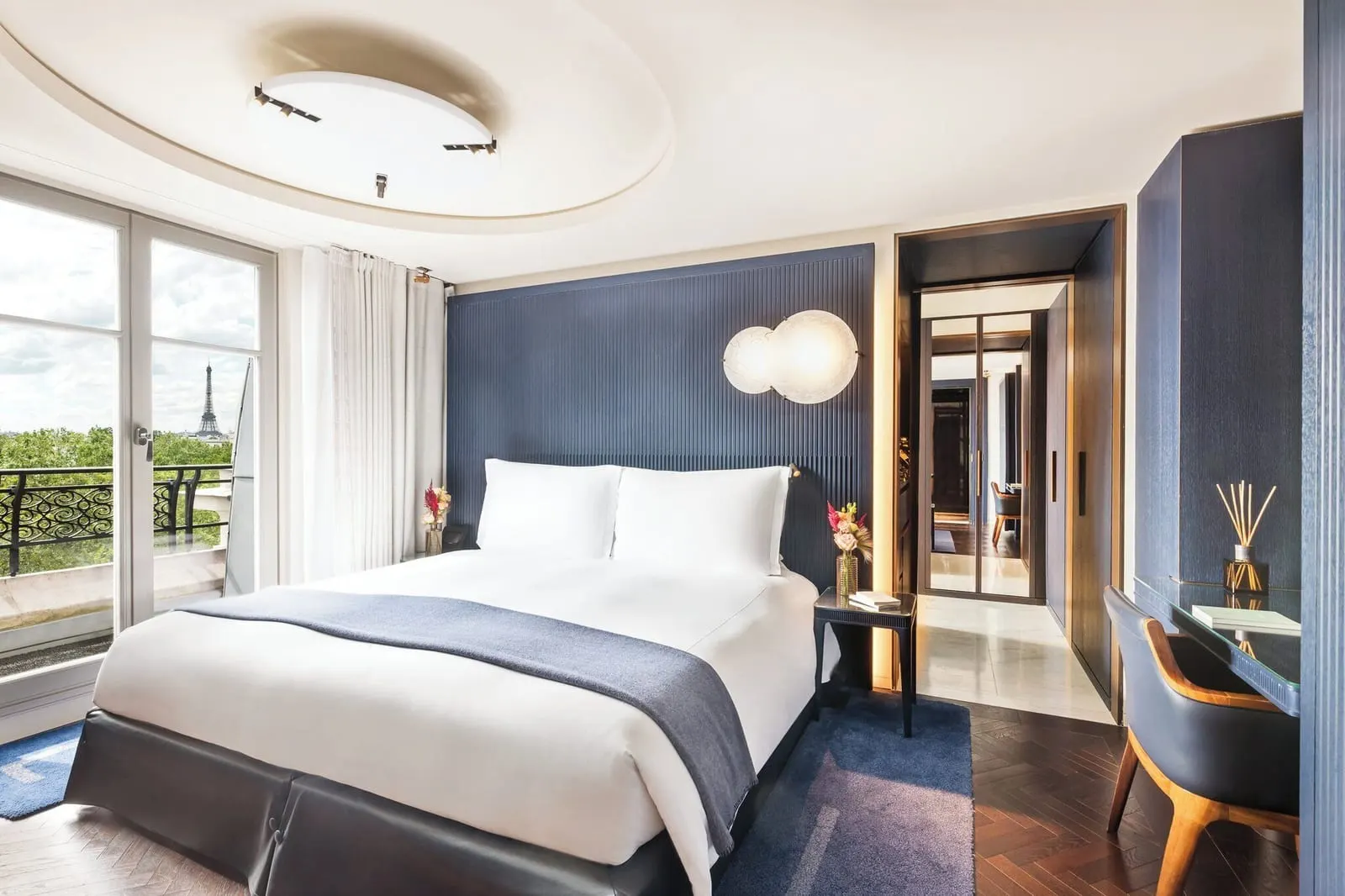 Two-Bedroom Eiffel Suite at Mandarin Oriental Lutetia, Paris: a double bed against a dark blue fluted headboard, a writing desk and French windows onto a wrought-iron balcony, with the Eiffel Tower beyond the trees