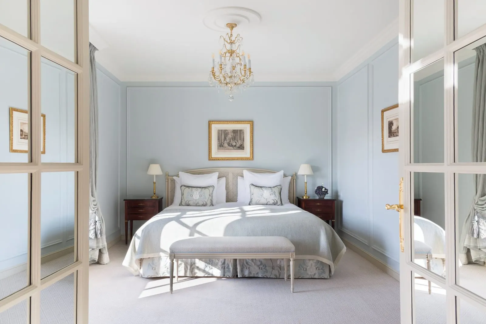 Deluxe Suite with balcony at Le Bristol Paris: the bedroom seen through glazed double doors, with a bed in pale grey linen, a bench at its foot and a crystal chandelier