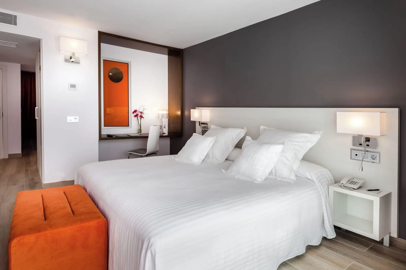 Suite at Barceló Corralejo Sands: a large bed with white linen, an orange footstool and a small desk with a mirror.