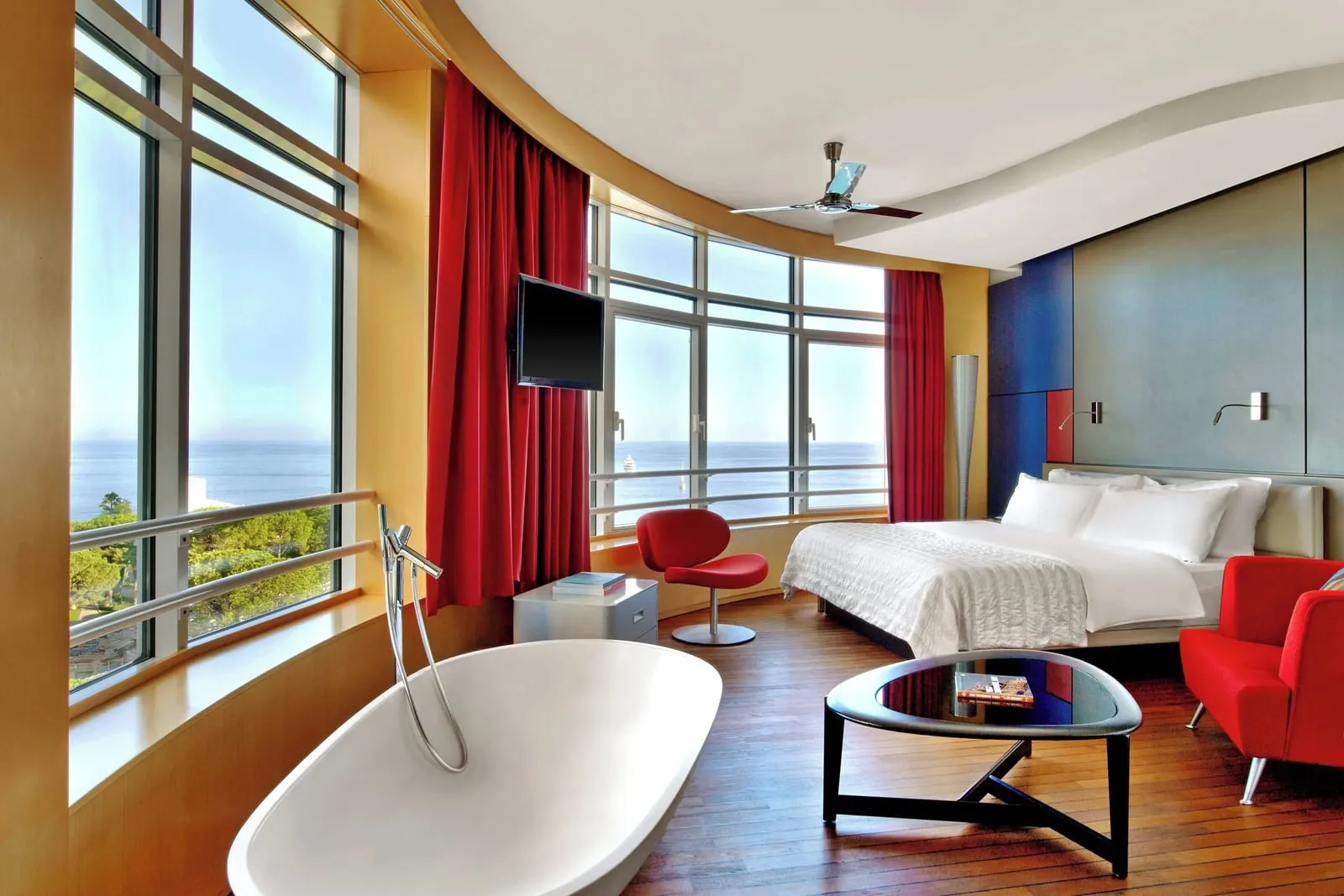 Design Suite at Le Méridien Beach Plaza: the bedroom with a freestanding bath, red chairs and curved windows onto the sea