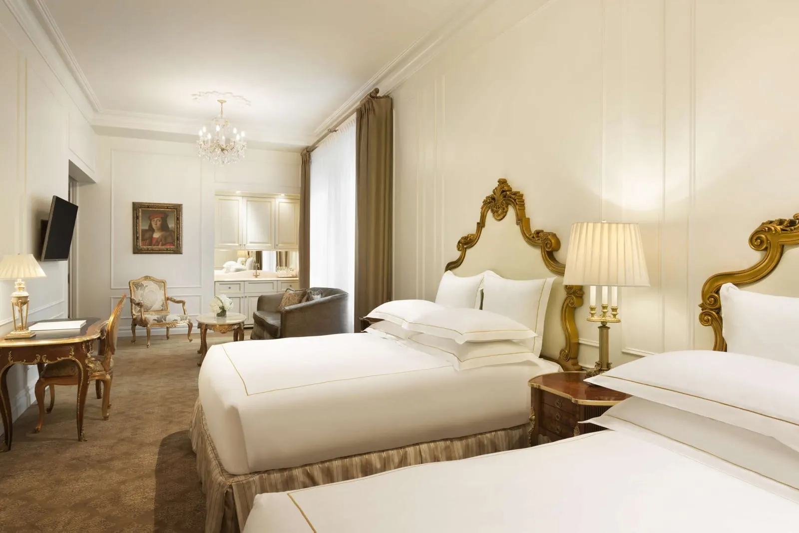 Family Grand Luxe Two Queens at The Plaza: two beds with gilded headboards, a desk, an armchair and a sofa beside a round table, and a bar area with glassware at the far end