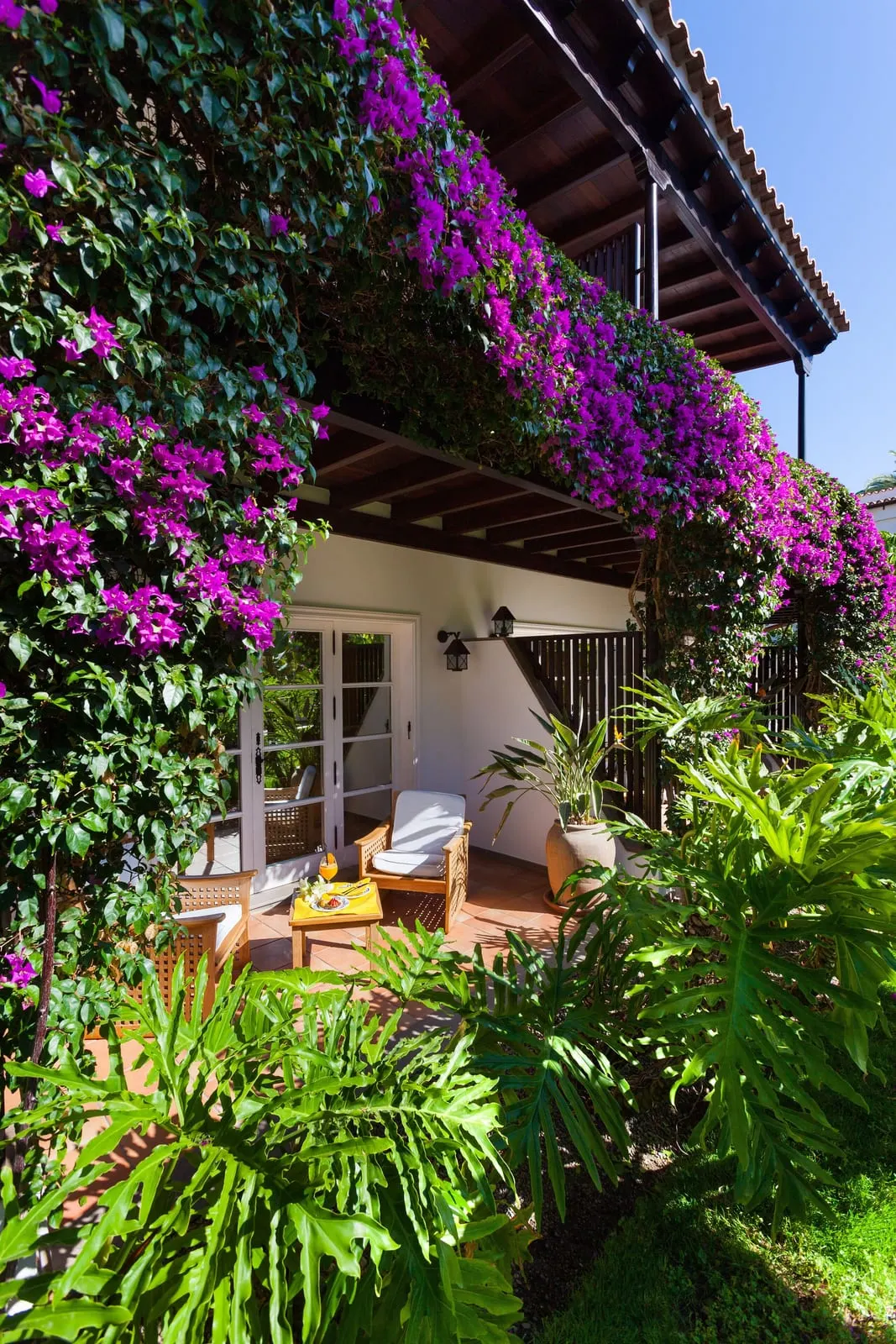 Superior Room at Seaside Grand Hotel Residencia: a terrace with teak armchairs and glazed doors, under a balcony hung with purple bougainvillea
