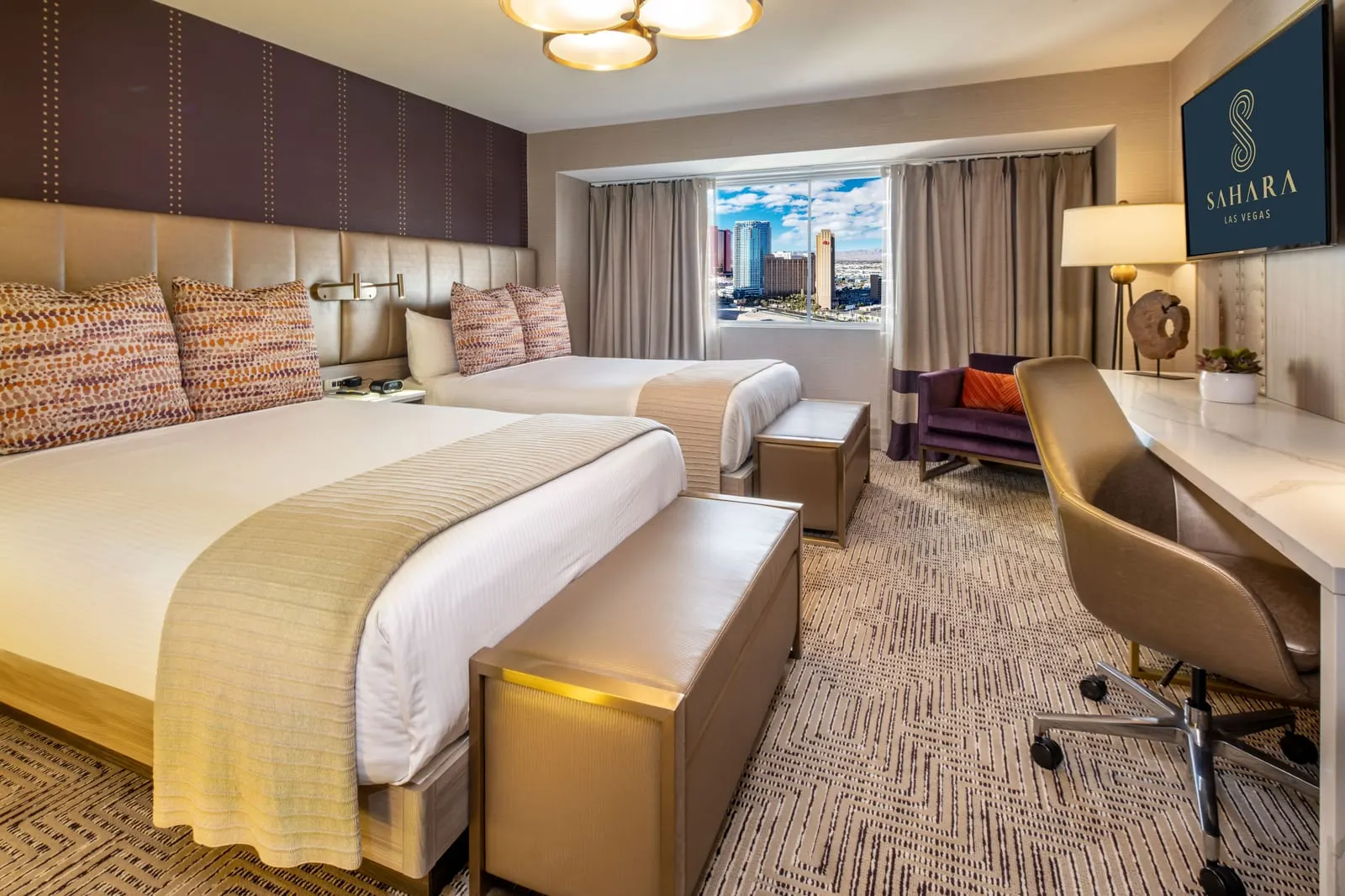 Marra Style Queens at SAHARA Las Vegas: two queen beds with padded headboards, a desk with an office chair, a purple armchair and a wall-mounted TV