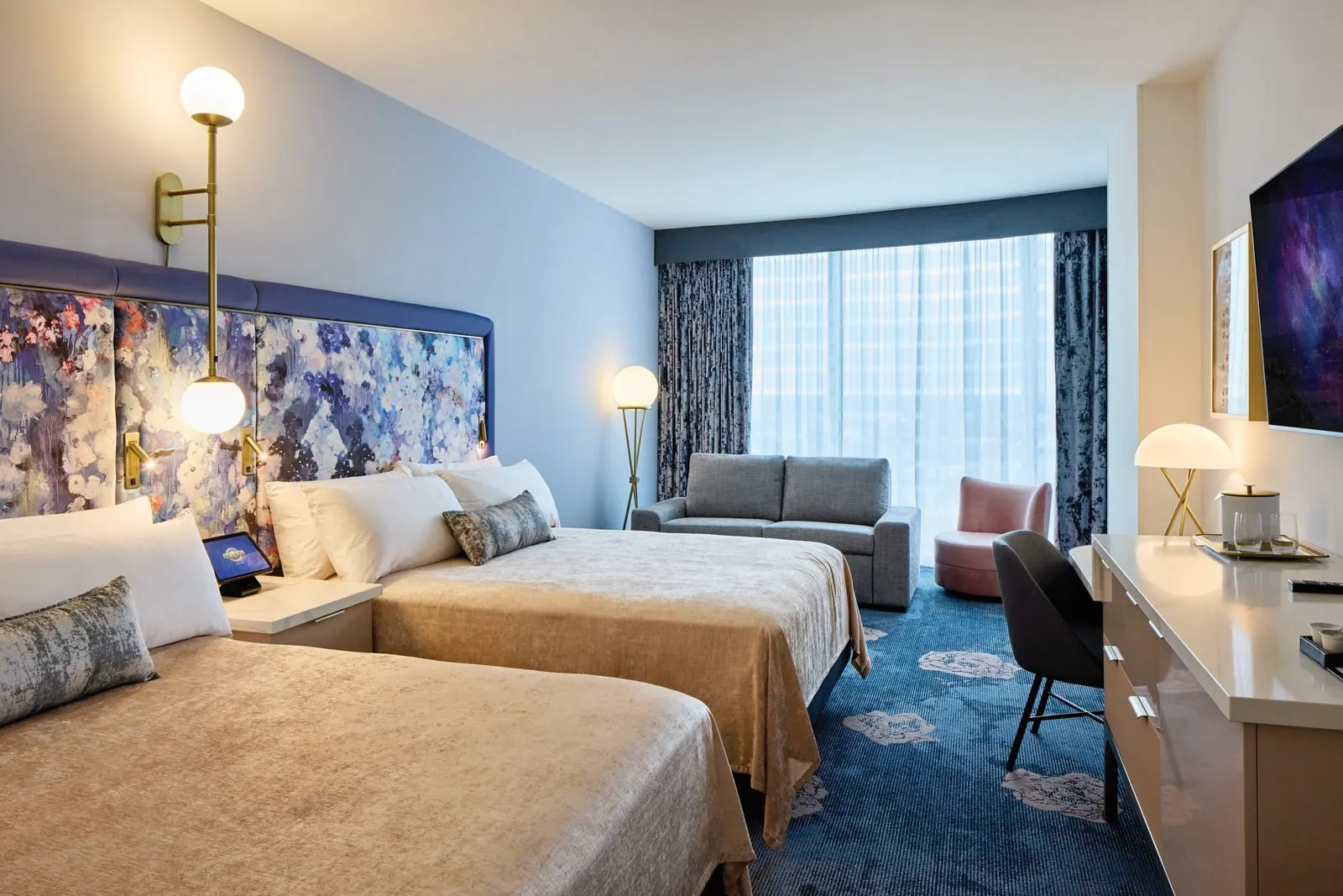 Deluxe 2 Queen Beds room at Universal Helios Grand Hotel: two queen beds with a floral headboard, a grey sofa and a pink armchair by the window, and a desk below a wall-mounted television