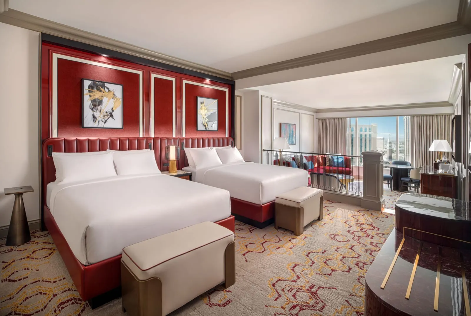 Newly Remodeled Luxury Two Queen Suite at The Venetian: two queen beds against a red panelled wall, a bench at the foot of each, and a railing beyond them above a living area with a red sofa
