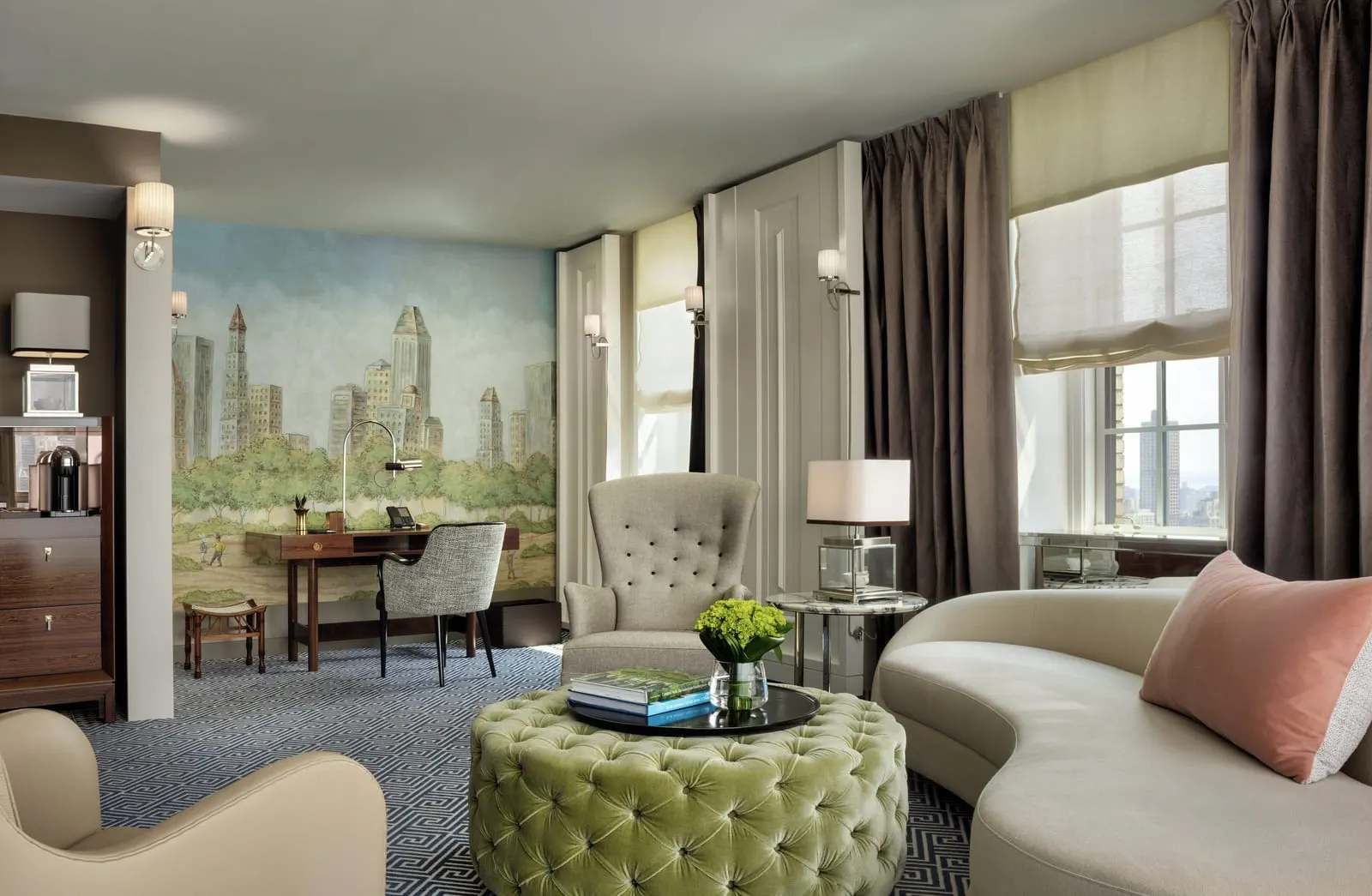 Rosewood Suite at The Carlyle: a living room with a curved cream sofa, a tufted green ottoman and a desk in front of a painted mural of trees and tall buildings