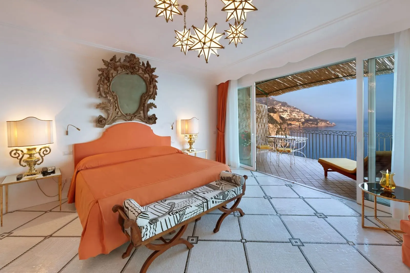 Prestige Room at Il San Pietro di Positano: a coral bed under a carved mirror, with open doors to a large shaded terrace over the sea and the coast
