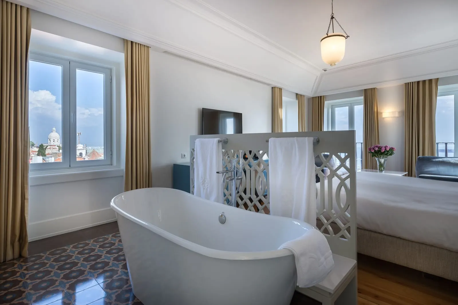 Santiago Suite at Santiago de Alfama: a freestanding bath by a window over the rooftops, with the bed beyond a lattice screen
